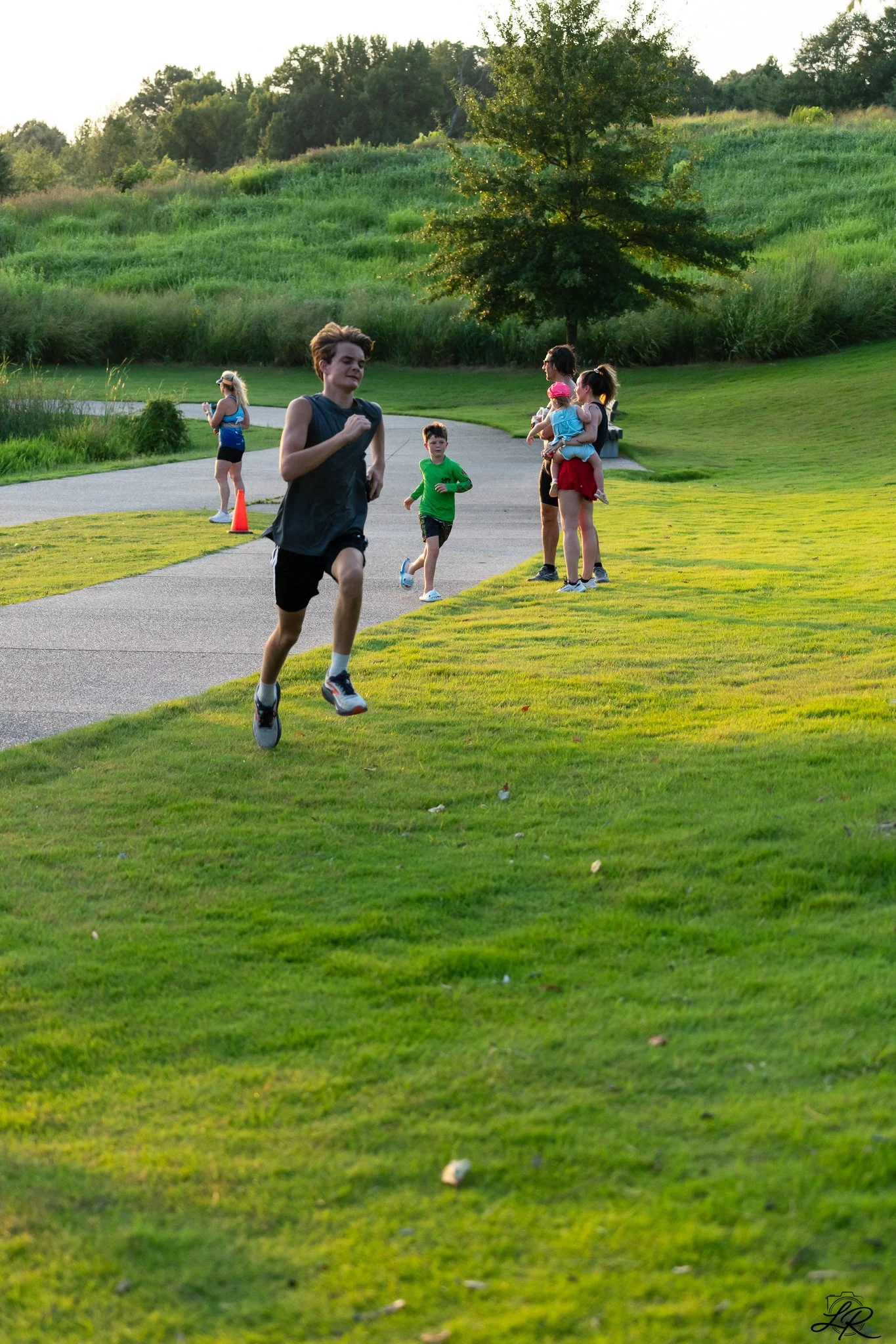 People jogging and walking in a park on a sunny day with a lush green landscape, trees, and a curved paved path.