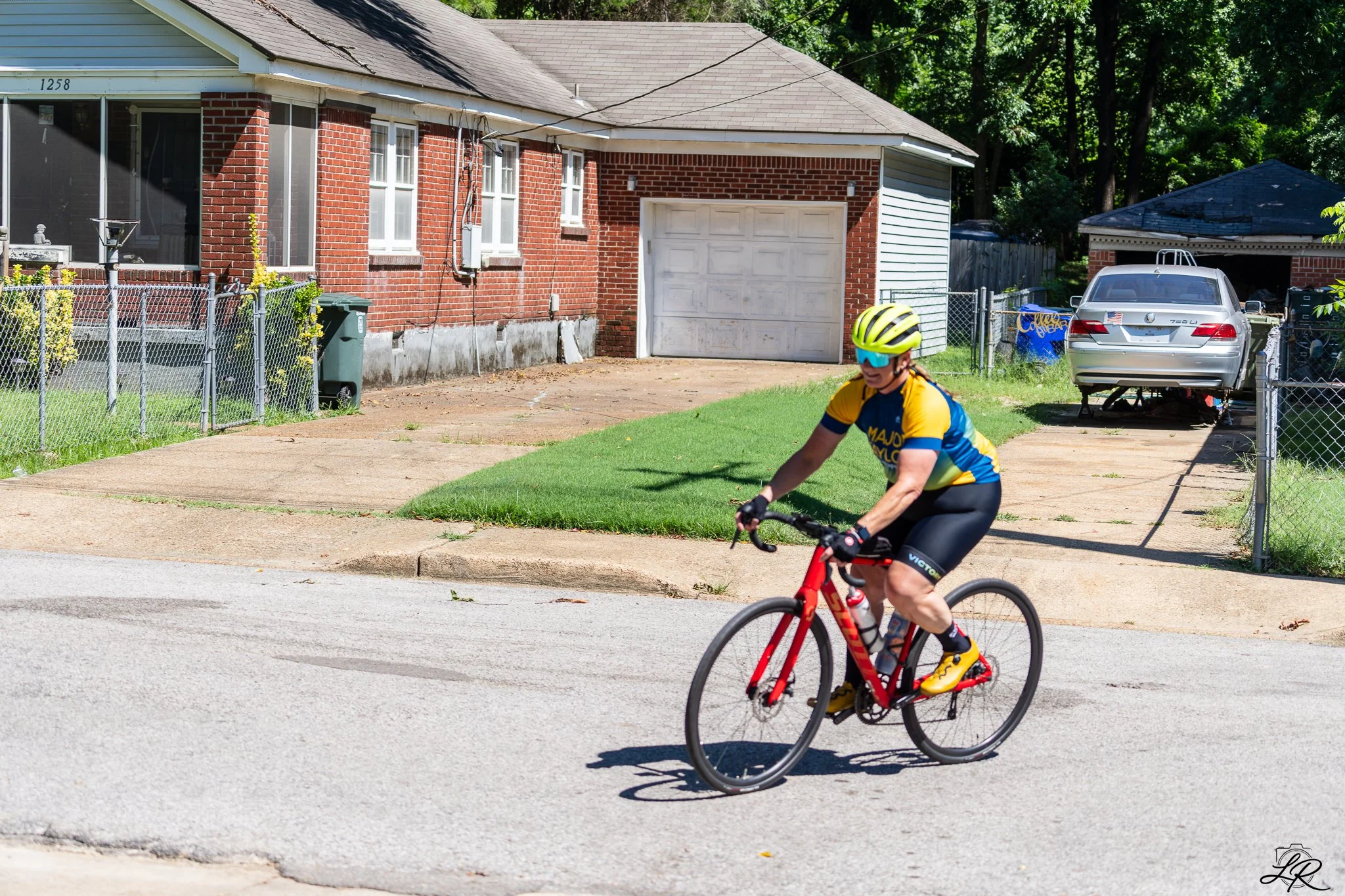 A young boy wearing a yellow helmet and sunglasses riding a red bicycle on a suburban street. In the background, there is a brick house with a front porch and a garage, a silver car parked in the driveway, and a lush green backyard with trees.