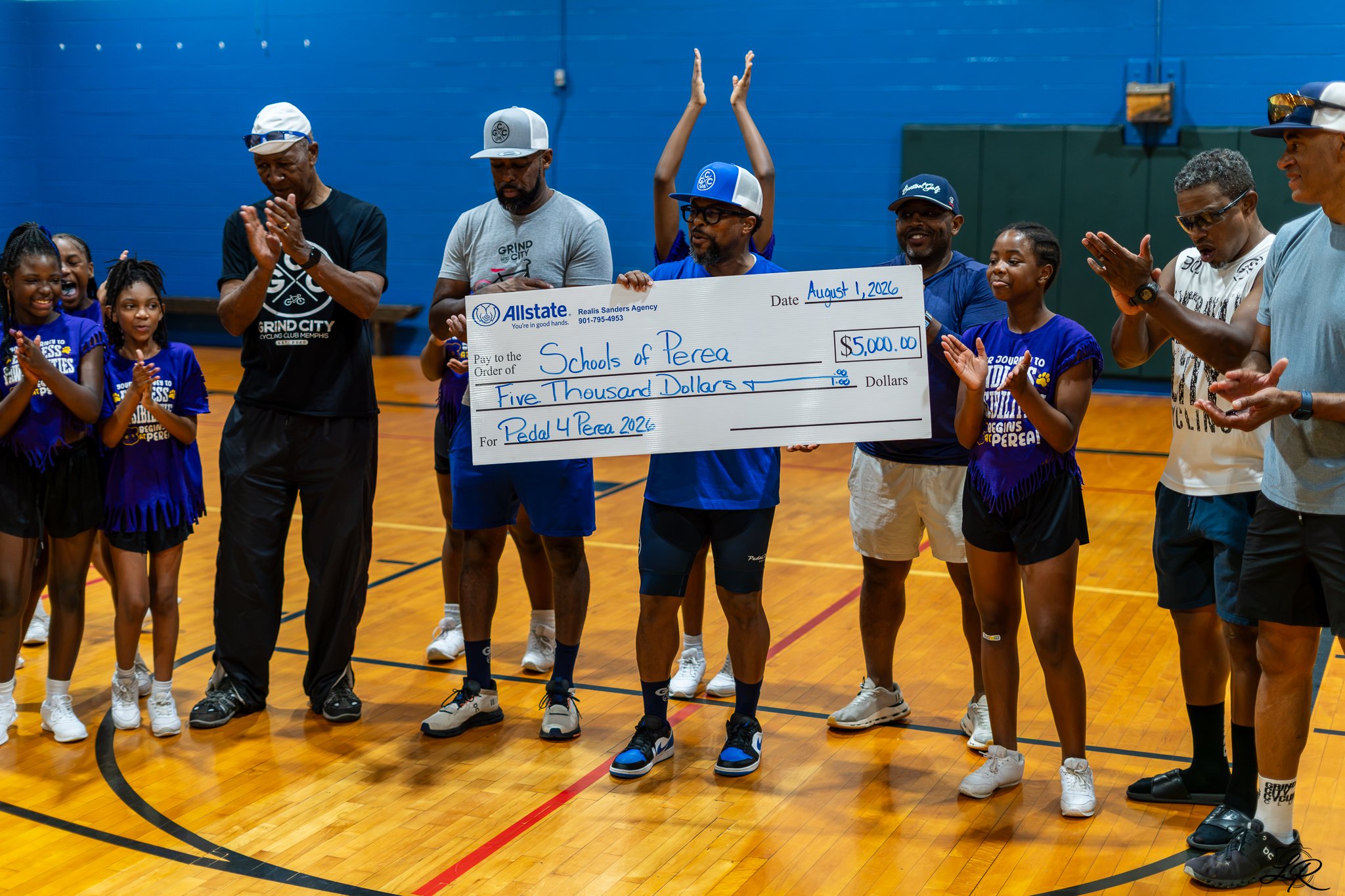 Group of children and adults gathered in a gymnasium, holding a large check for $5,000 made out to Schools of Perea. The date on the check is August 1, 2026, celebrating a donation for Pedal 4 Perea 2026. Some people are clapping, and others are rais