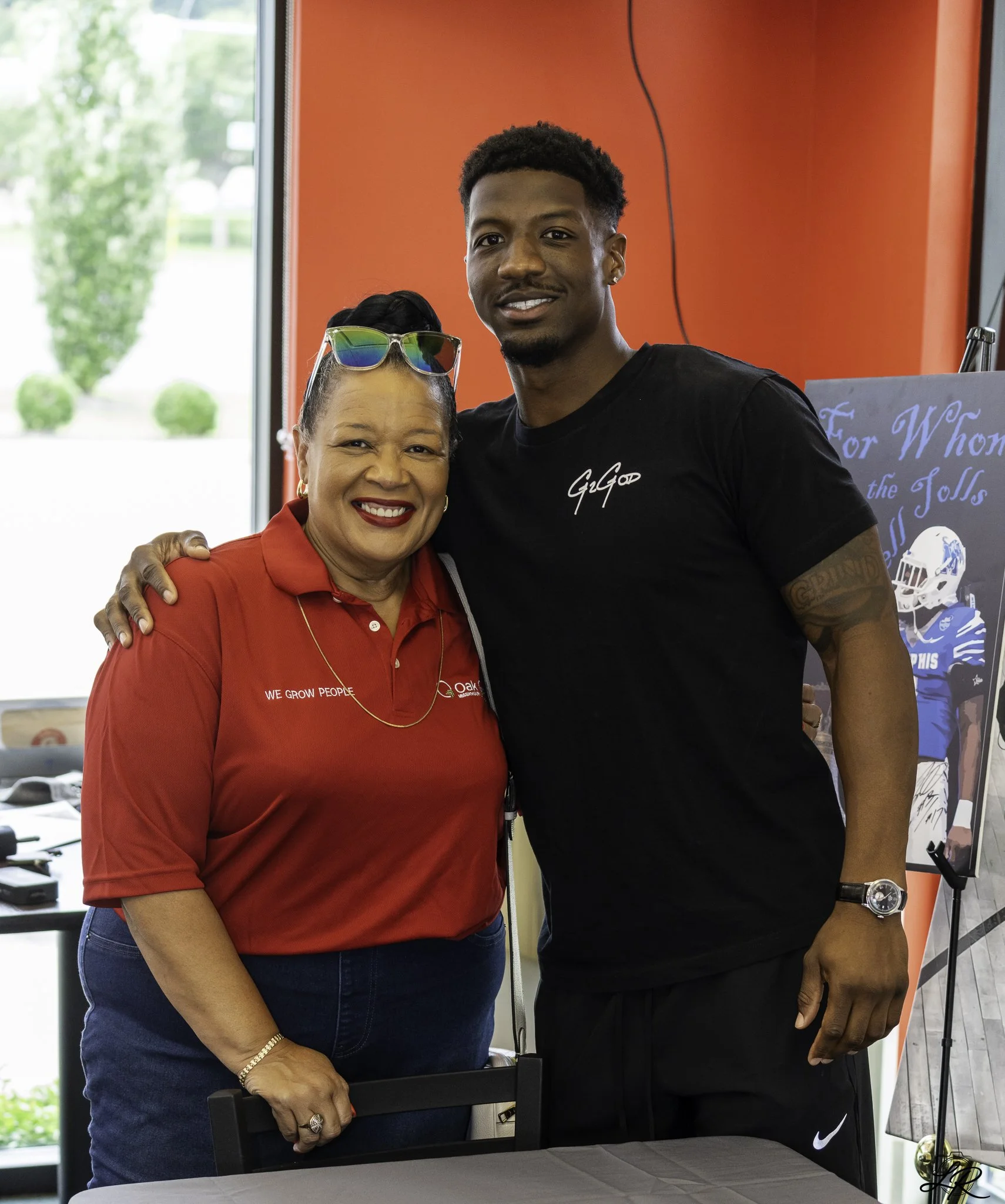 A woman and a man standing together indoors, smiling at the camera. The woman, wearing a red shirt with the words 'WE GROW PEOPLE' and glasses on her head, has her arm around the man. The man is wearing a black T-shirt and a watch, and has his arm ar