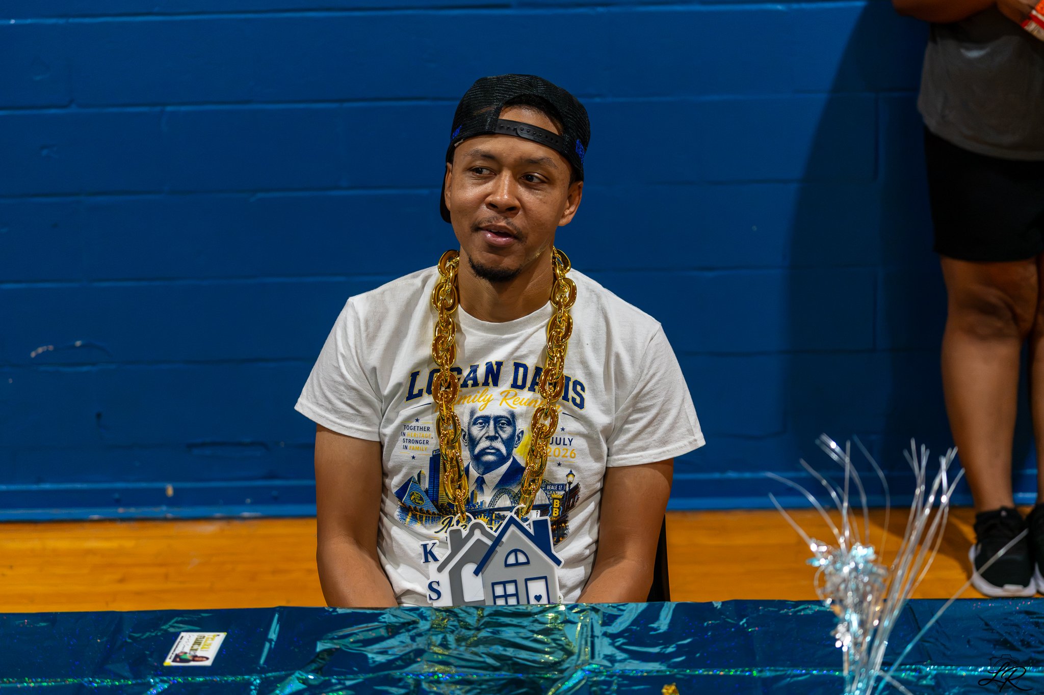 A man wearing a black cap backwards, a gold chain, and a white T-shirt sitting at a table decorated with shiny blue wrapping paper. The T-shirt features a graphic of a man in a suit and the words "LOS ANGELES" and "Family Reunion." Behind him is a bl
