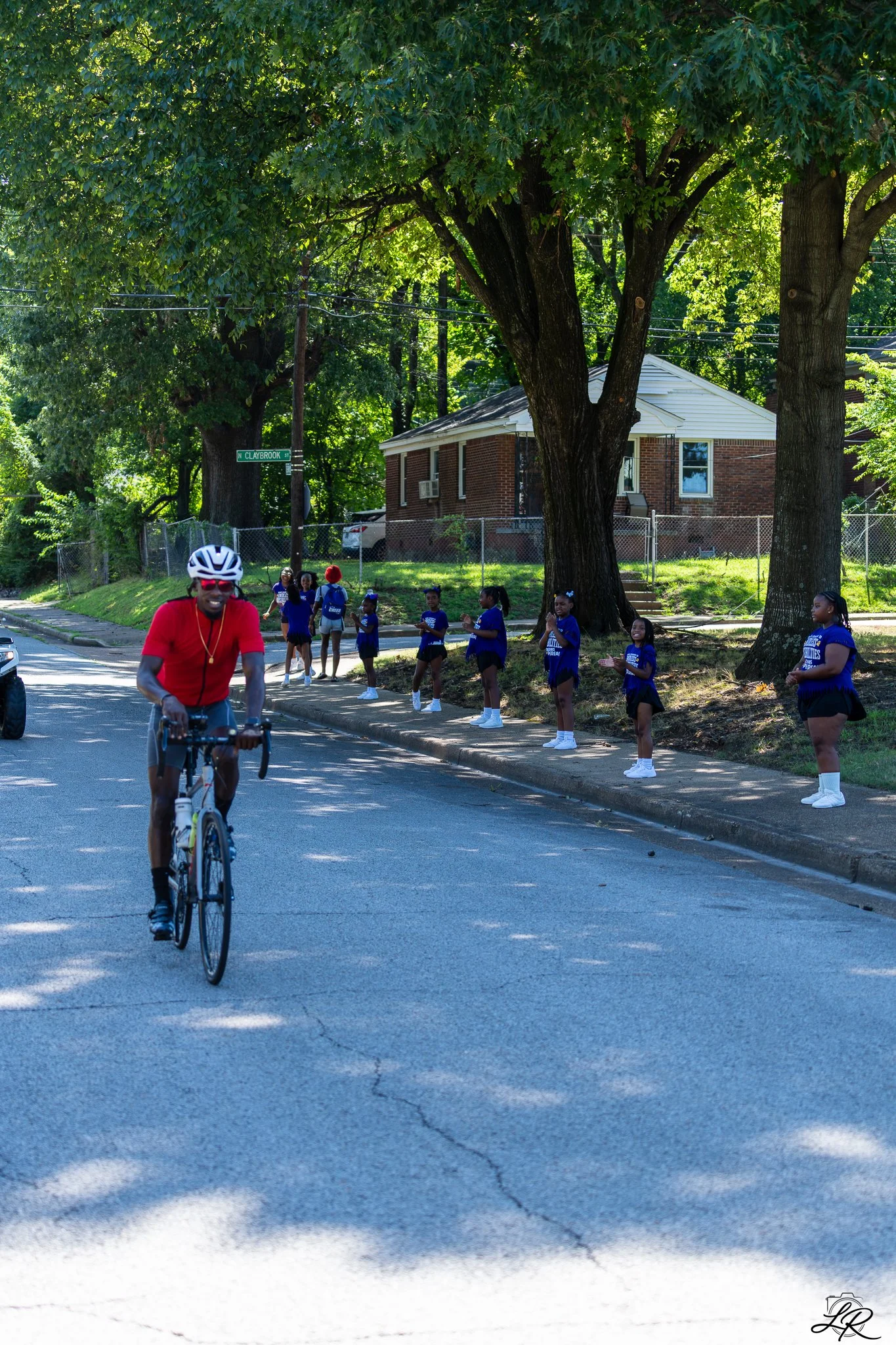 A man riding a bicycle on a sunny residential street, with a group of children standing and clapping on the sidewalk, and large trees providing shade.