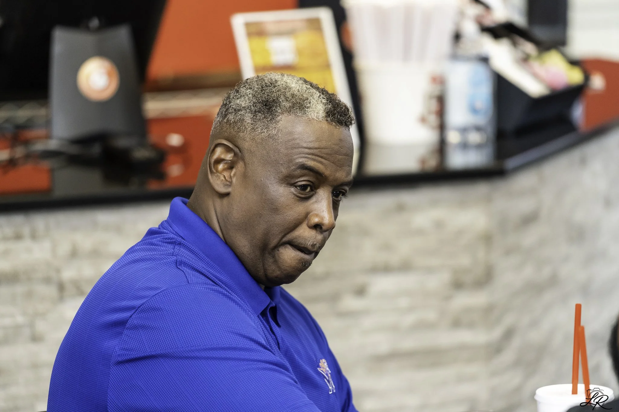 A middle-aged man with gray, curly hair and a dark skin tone wearing a blue shirt, seated at a counter in a casual setting, possibly a cafe or restaurant, with blurred background including a red wall, a menu board, and a drink with a red straw.