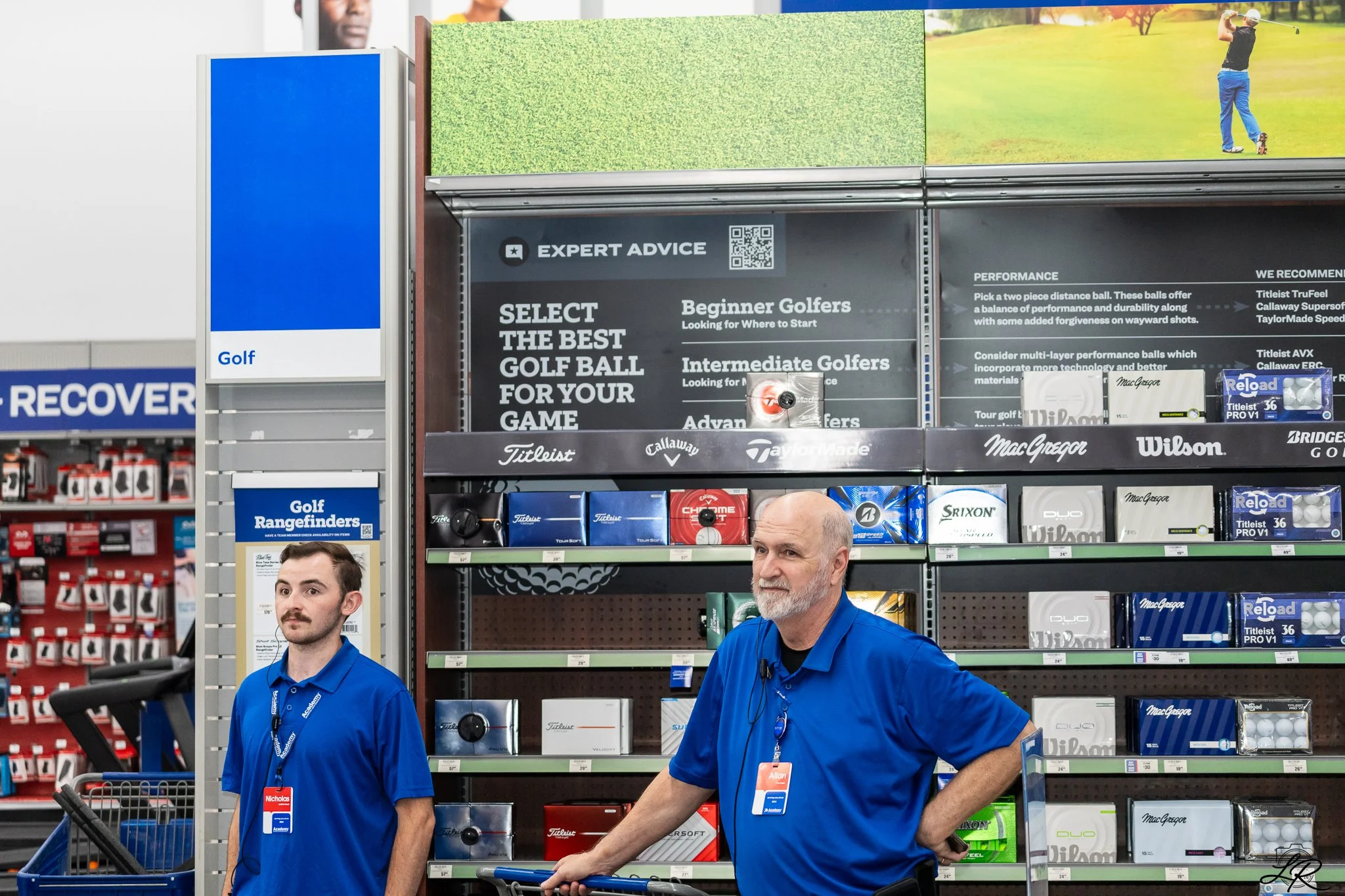Two employees wearing blue shirts work at a golf equipment display in a store. Behind them, there are shelves and signage related to golf balls and accessories, with a large promotional sign above. The store environment is brightly lit, with other pr