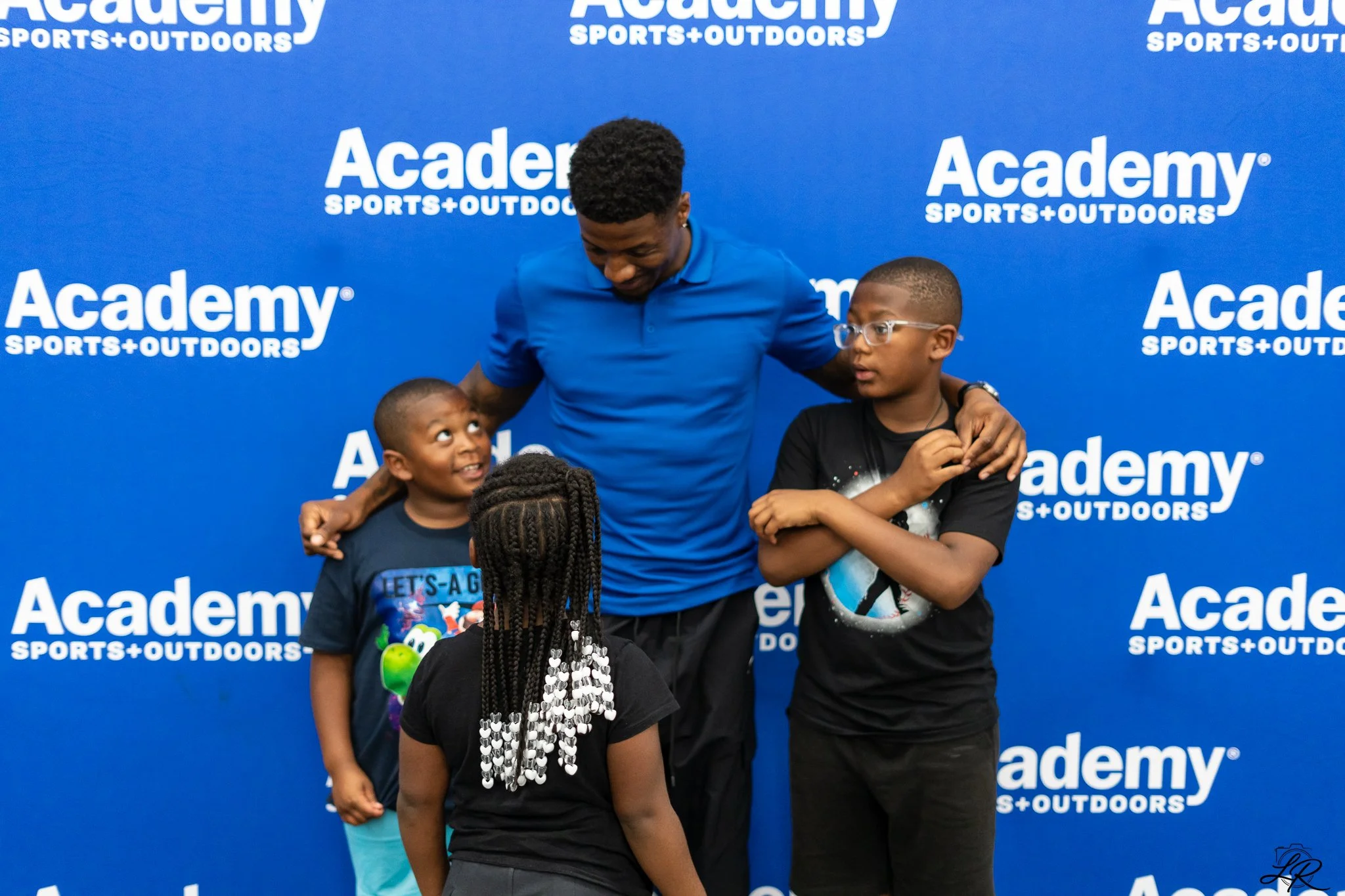 A man embraces two young boys in front of a blue backdrop that reads 'Academy Sports + Outdoors' repeatedly. The man looks down smiling, the boys are engaged in conversation, one boy has glasses and the other has hair styled in braids with white bead