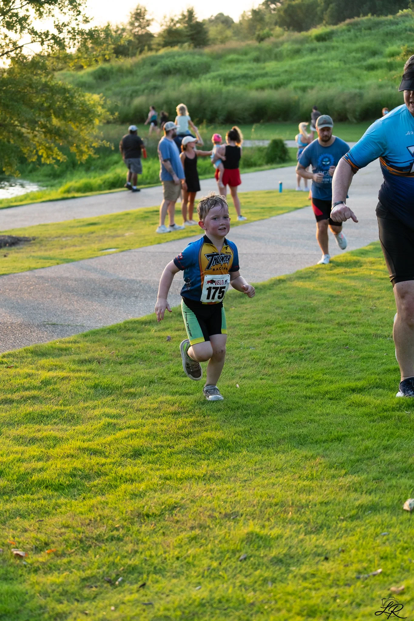 A boy running in a race on a grassy field with people watching and walking along a path in the background, trees, and a hill in the distance during the daytime.