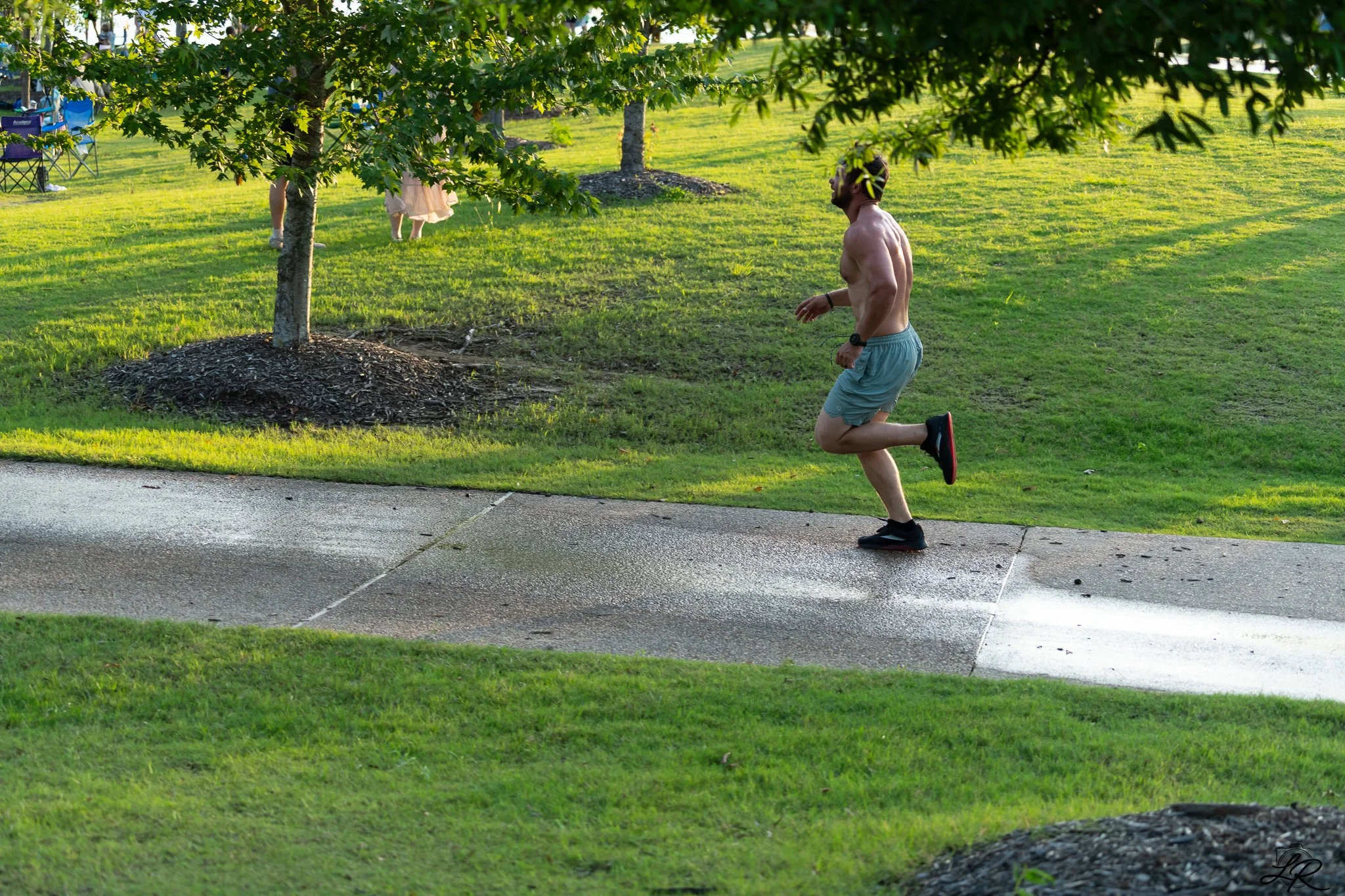 A shirtless man running on a sidewalk in a park with green grass, trees, and some people in the background.