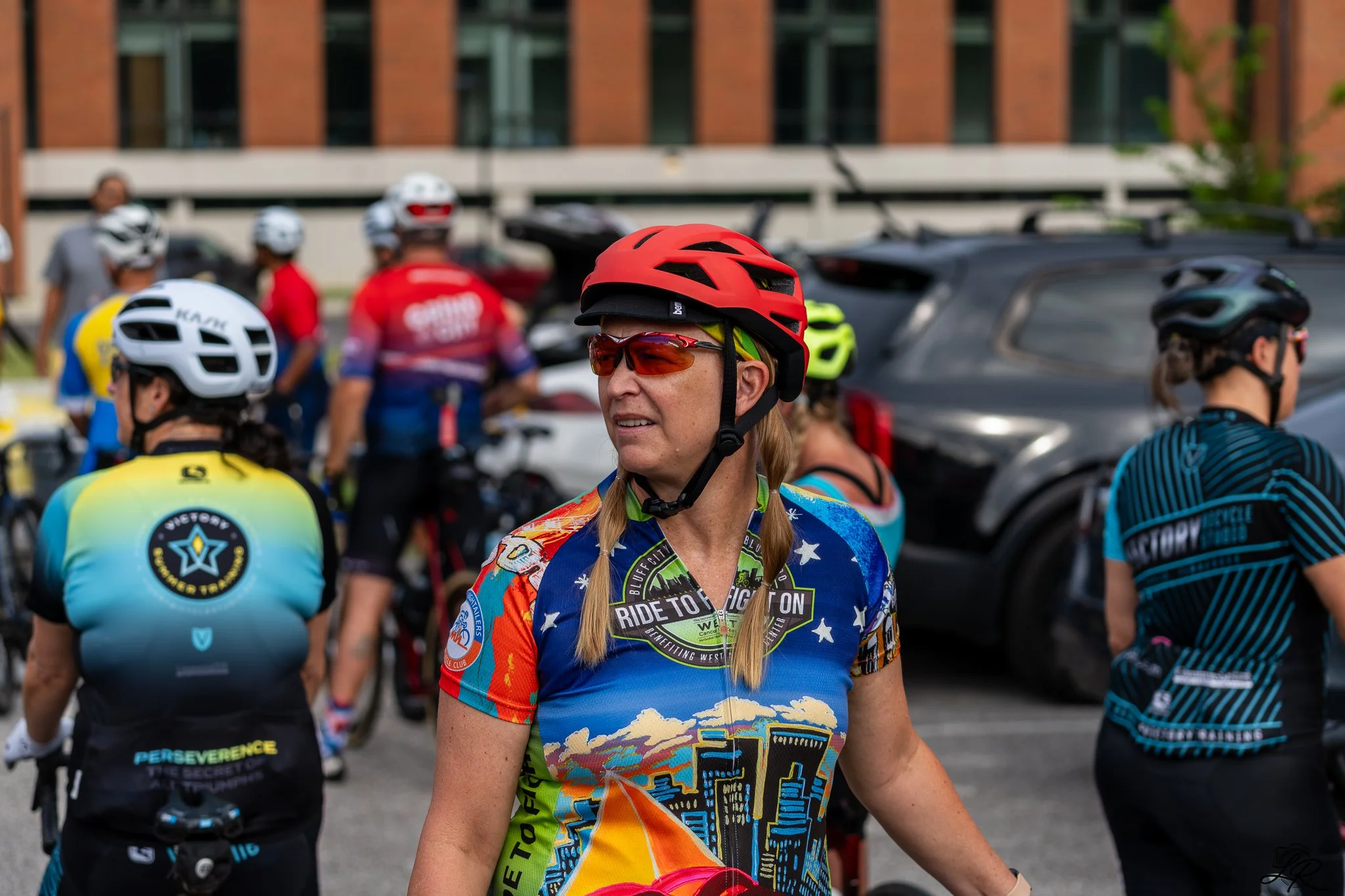 A group of cyclists gathering outdoors, with a woman in a colorful cycling jersey, red helmet, and sunglasses in the foreground.