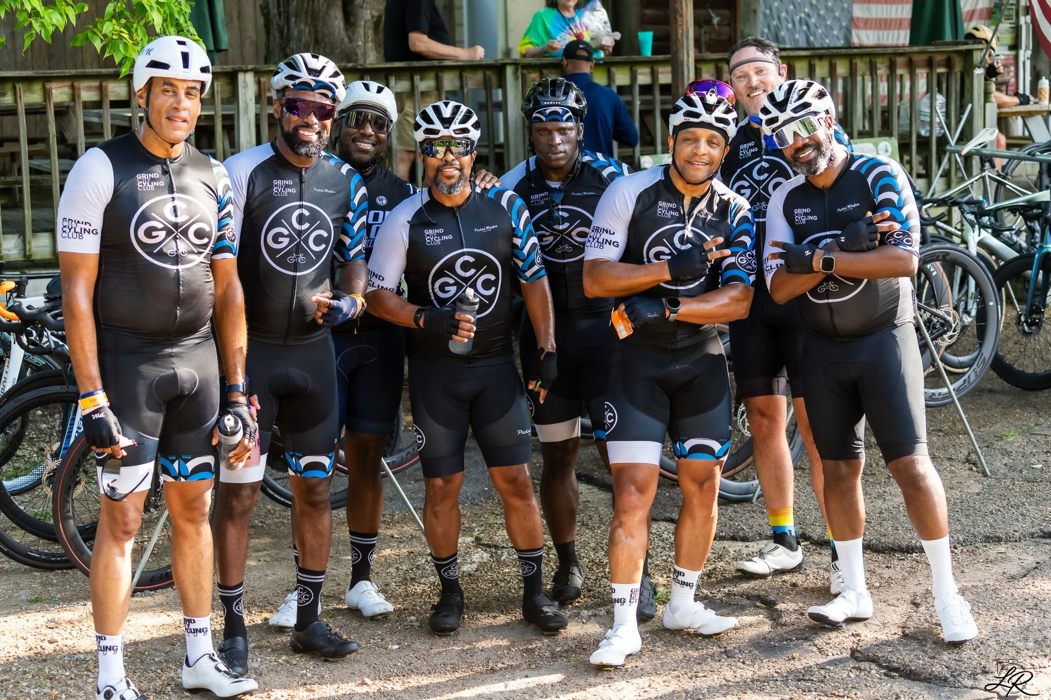 A group of eight diverse male cyclists in matching black, white, and blue cycling jerseys and shorts, wearing helmets and sunglasses, standing outdoors with bicycles visible in the background. They are smiling and posing for the photo.