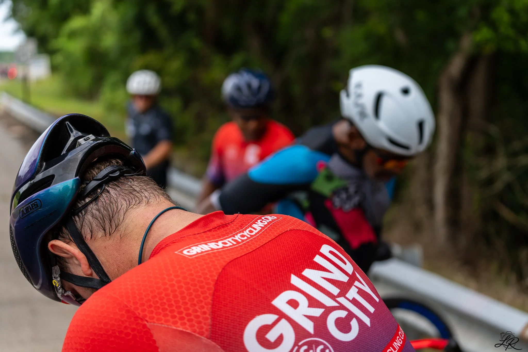 Close-up of a cyclist wearing a red jersey and a blue helmet, with other cyclists in helmets and colorful jerseys in the background, on a tree-lined road.