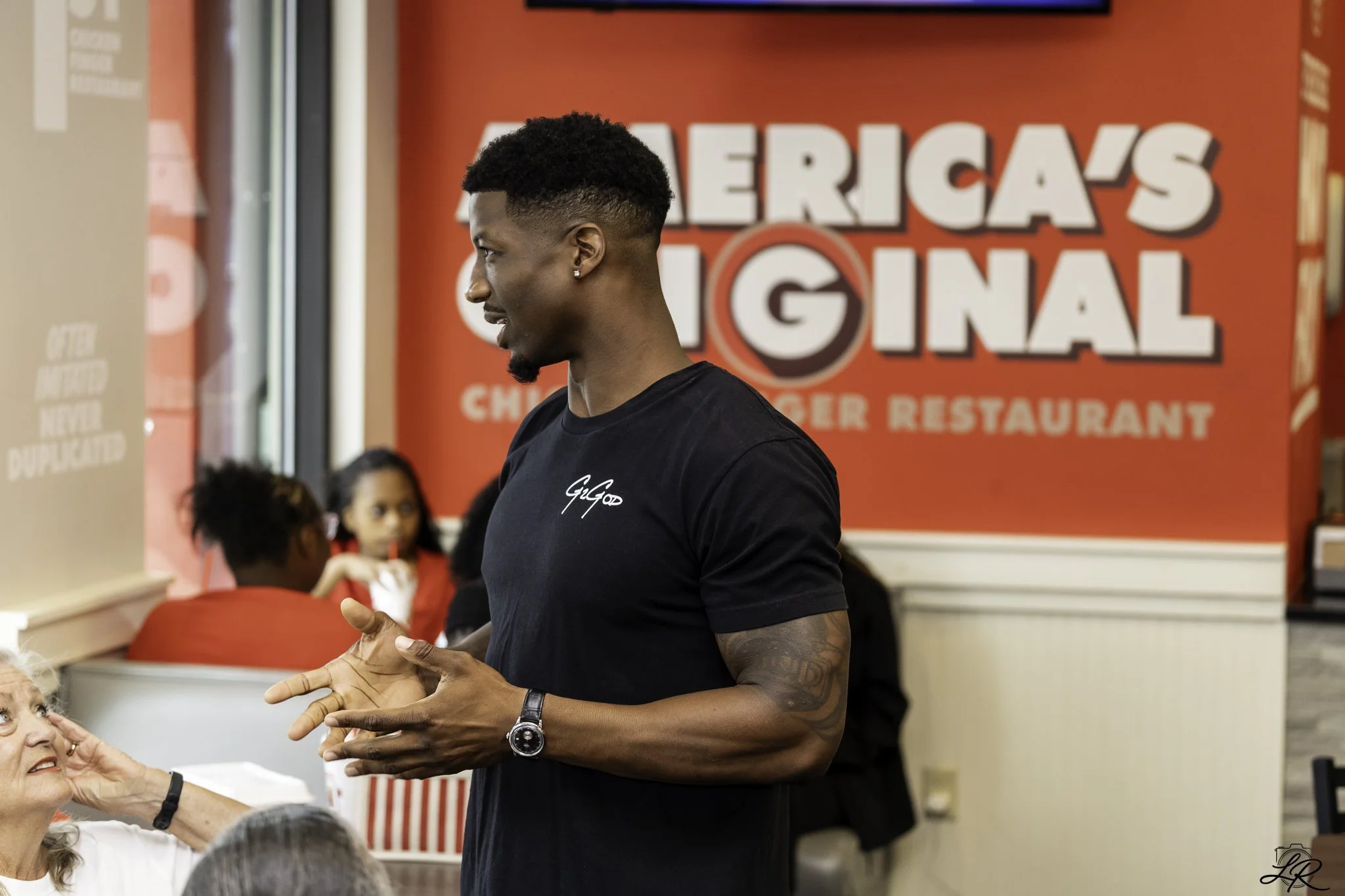 A young man with short curly hair, earrings, and a black T-shirt is talking to an elderly woman with gray hair, who is sitting at a table inside a restaurant. The restaurant has a red background with large white lettering that reads 'AMERICAS ORIGINA