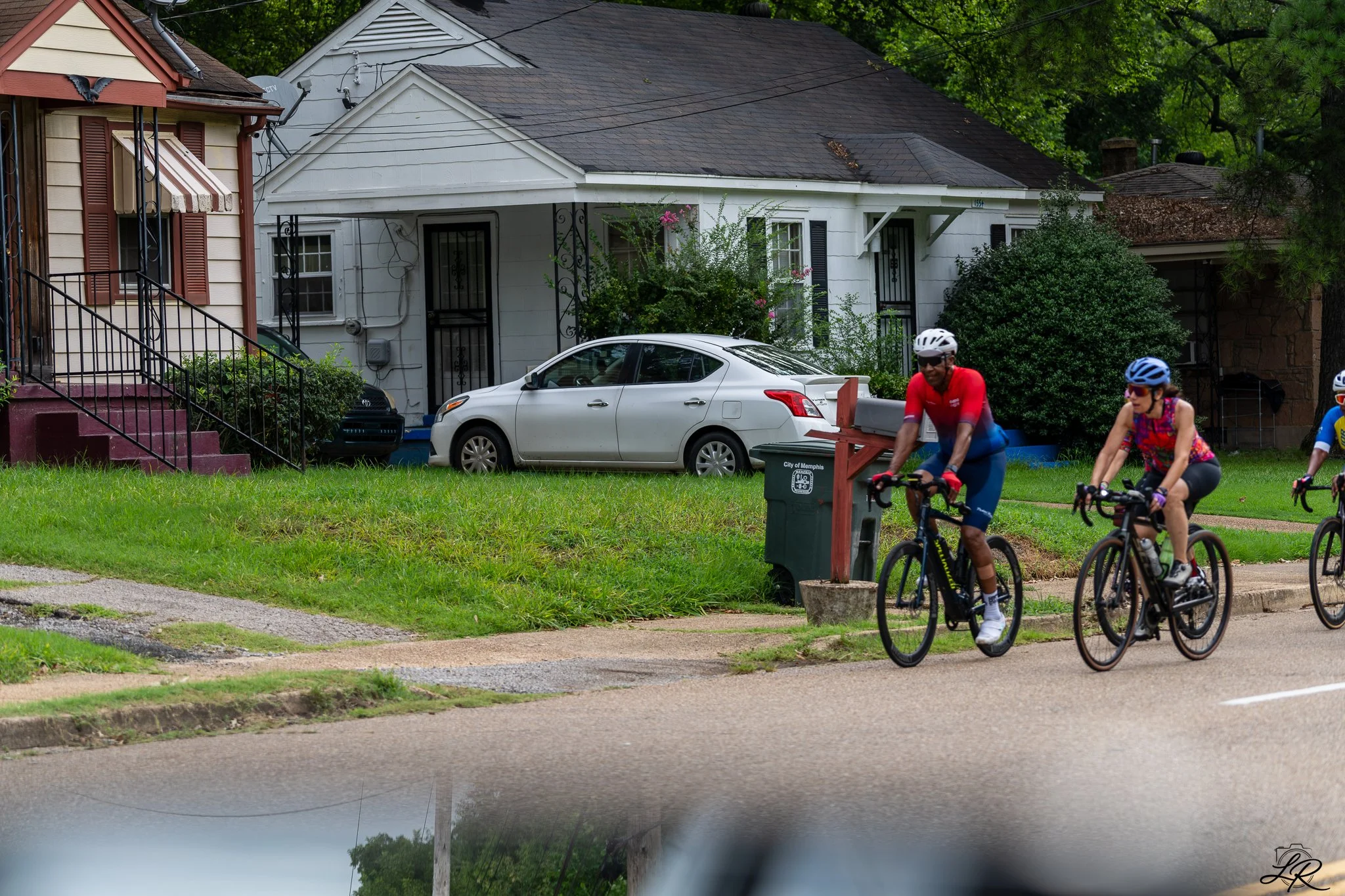 A group of people riding bicycles on a sidewalk in a residential neighborhood with houses, cars, and greenery in the background.