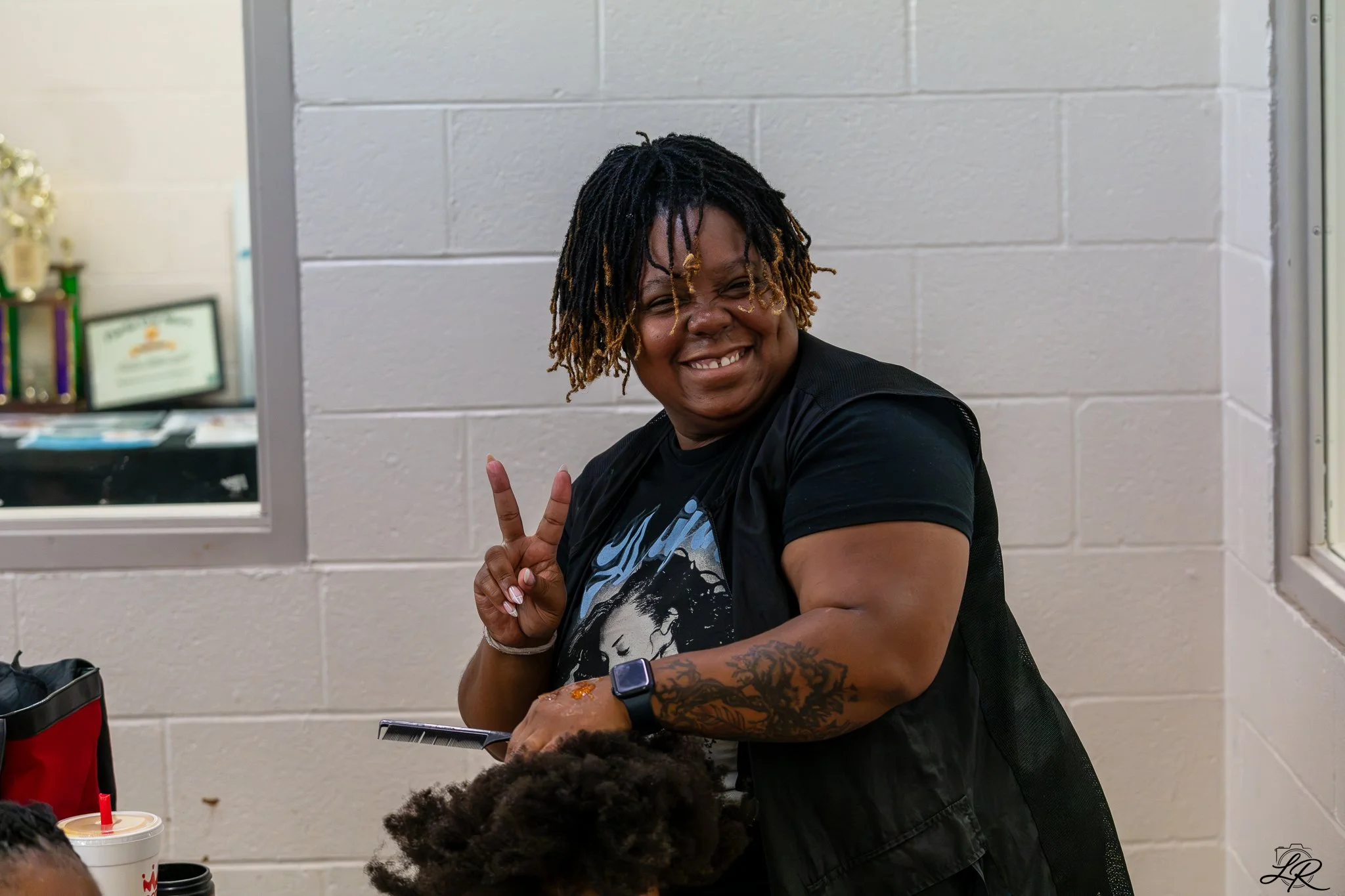 A smiling woman with dreadlocks making a peace sign with her fingers inside a room with a white brick wall. On the table in front of her, there are drinks, and in the background, there are trophies and framed certificates.