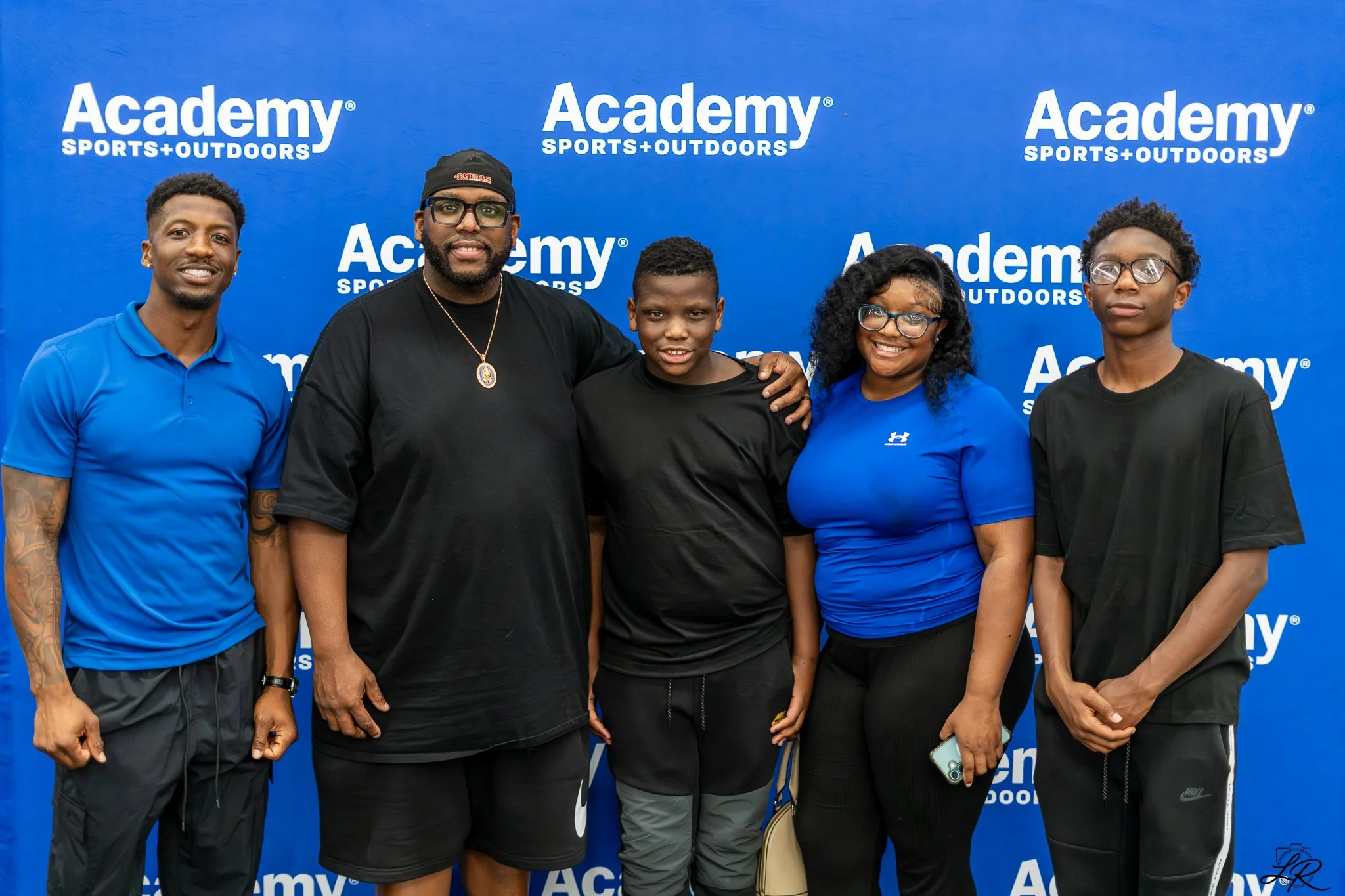 Group of five people standing in front of a blue banner with 'Academy Sports + Outdoors' written on it, posing for a photo.