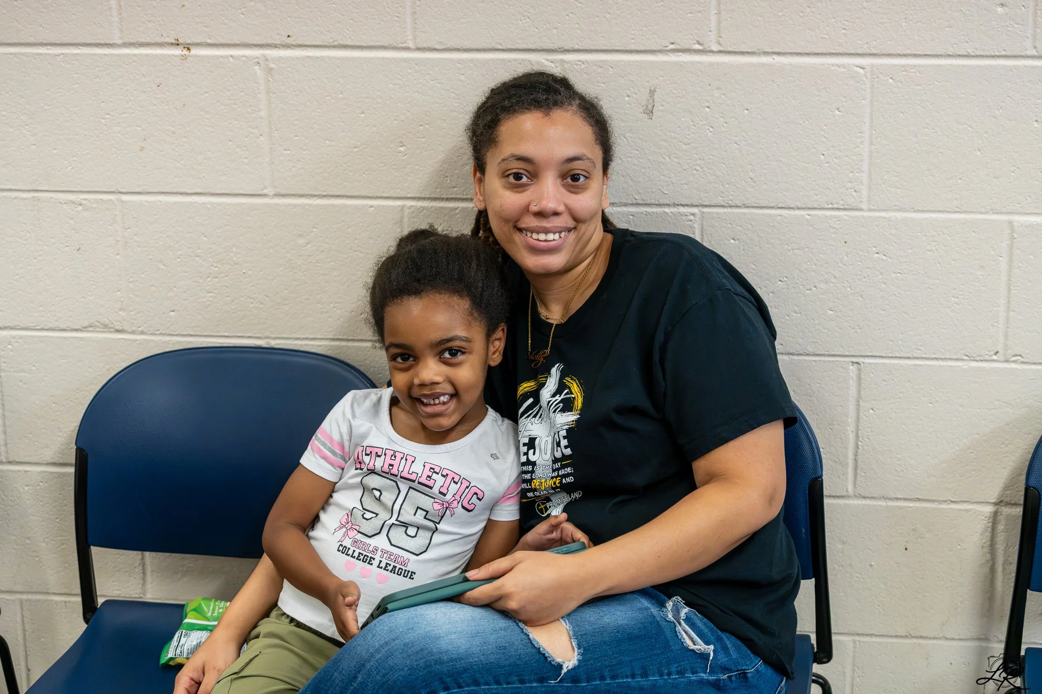 A woman and a young girl sitting together, smiling, against a beige brick wall. The woman has dark curly hair pulled back, a nose piercing, and is wearing a black T-shirt with gold and white text. The girl has dark hair in a ponytail, is wearing a wh