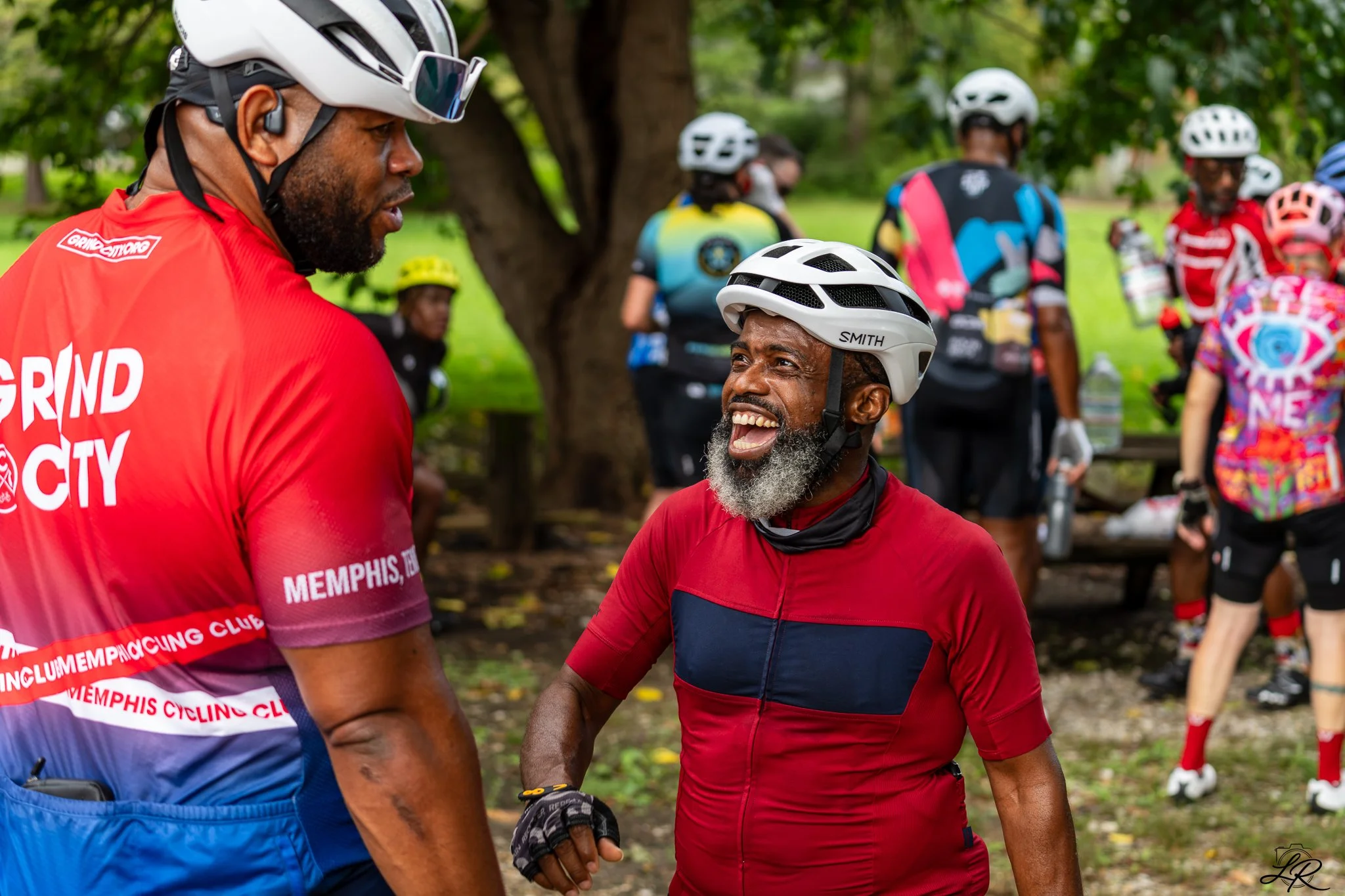 Two men wearing cycling helmets and red cycling jerseys sharing a moment of joy while surrounded by other cyclists in a park.