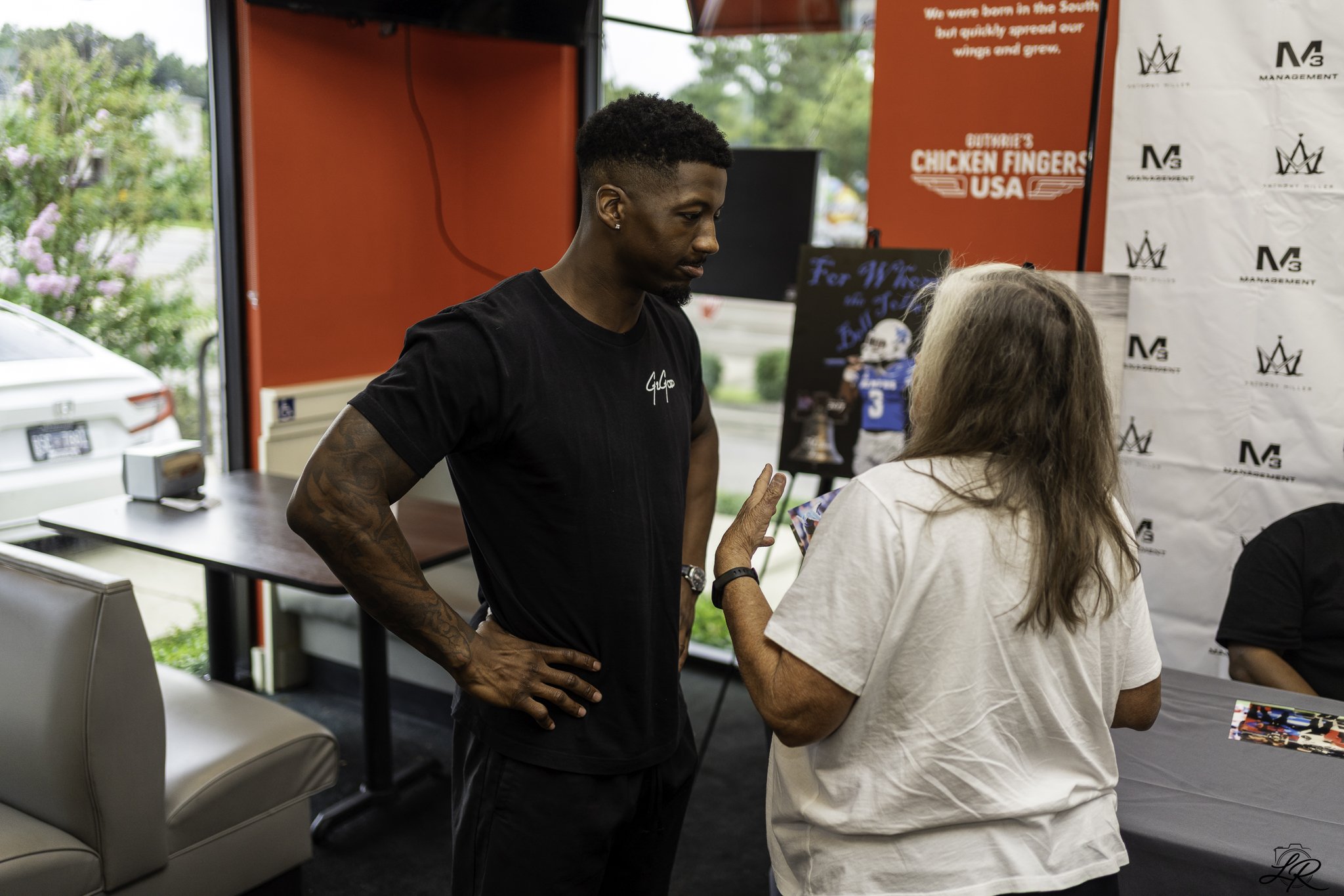 A young man with dark hair and tattoos on his arm is engaged in conversation with an older woman with gray hair inside a red and white booth at an event. There are promotional materials and banners behind them, including one reading 'Chicken Fingers 
