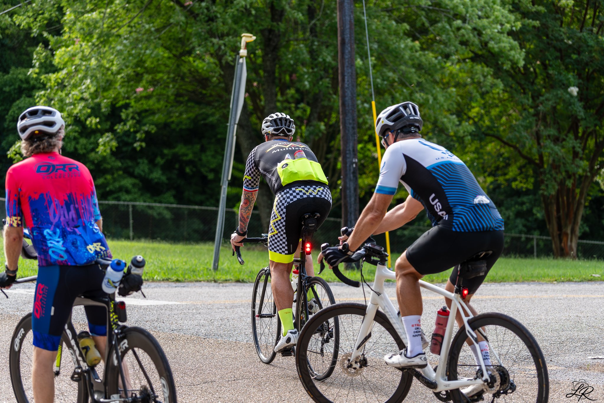 Group of cyclists wearing helmets and colorful cycling attire, gathered outdoors near a wooded area, with some on bikes and others standing.
