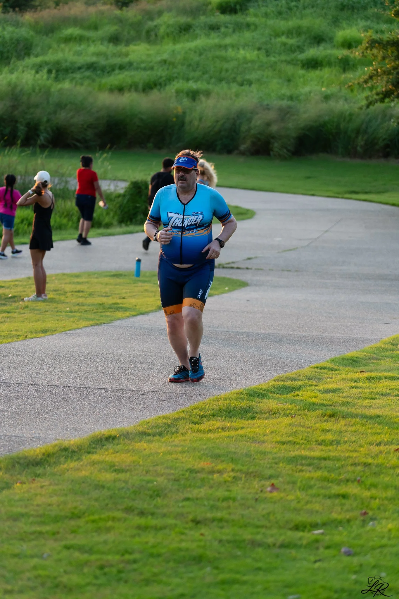 A man running on a paved path in a park with green grass and shrubs, wearing a blue athletic outfit and visor.