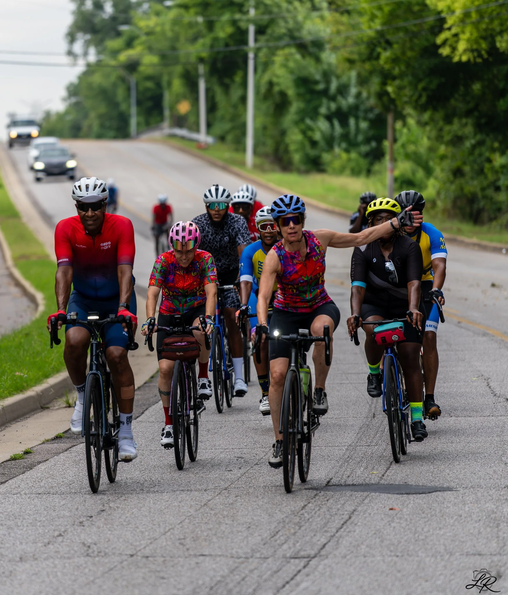 A group of cyclists wearing helmets riding on a suburban road surrounded by green trees and utility poles.