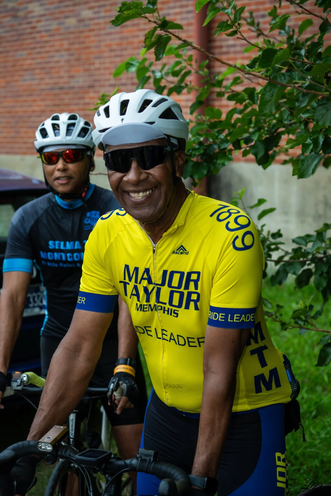 Two cyclists wearing helmets and sunglasses, smiling and posing for a photo outdoors near greenery, with a brick wall in the background. One cyclist is wearing a yellow jersey with 