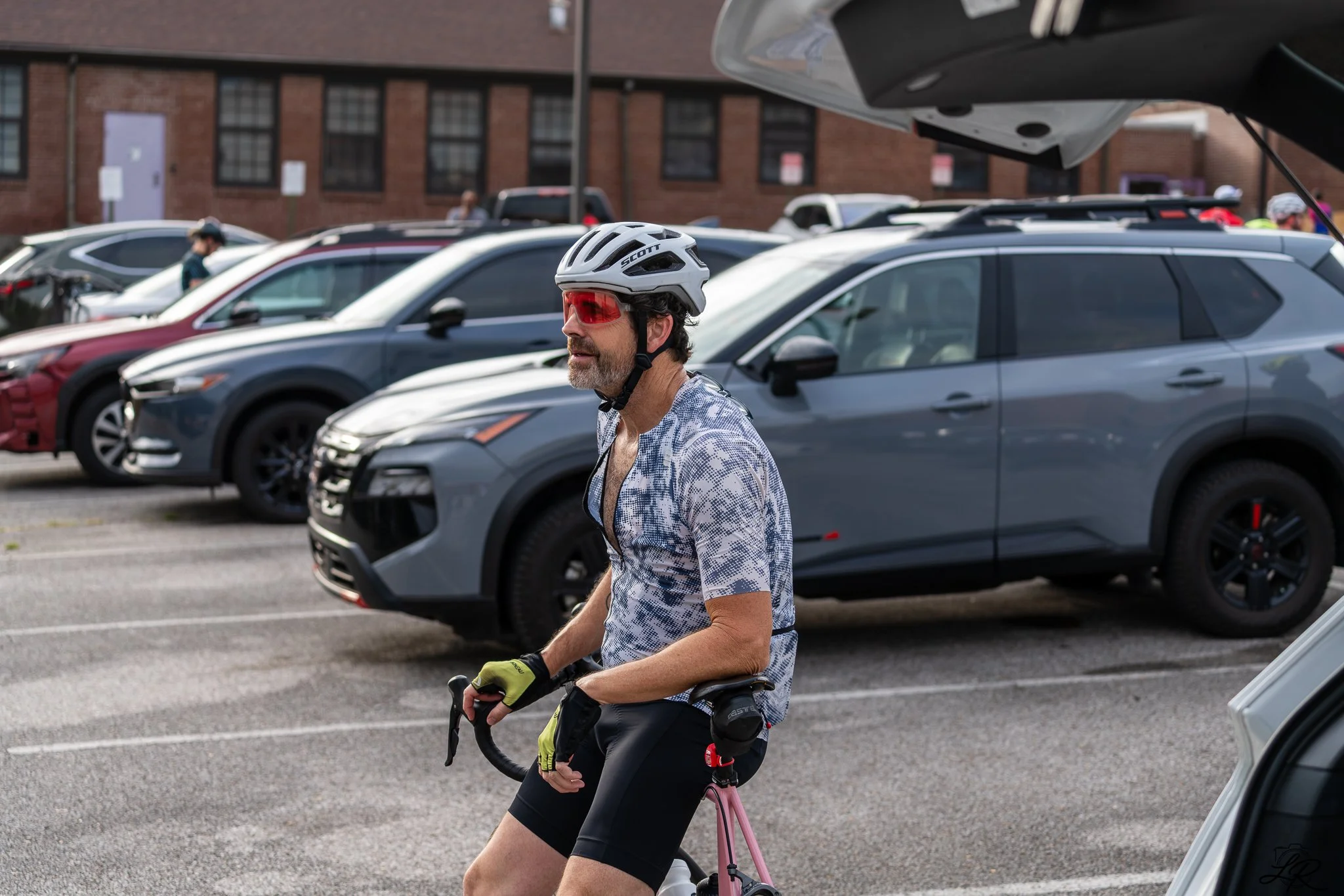 A man wearing a white bike helmet, red sunglasses, and a patterned shirt riding a pink bicycle in a parking lot filled with parked cars.