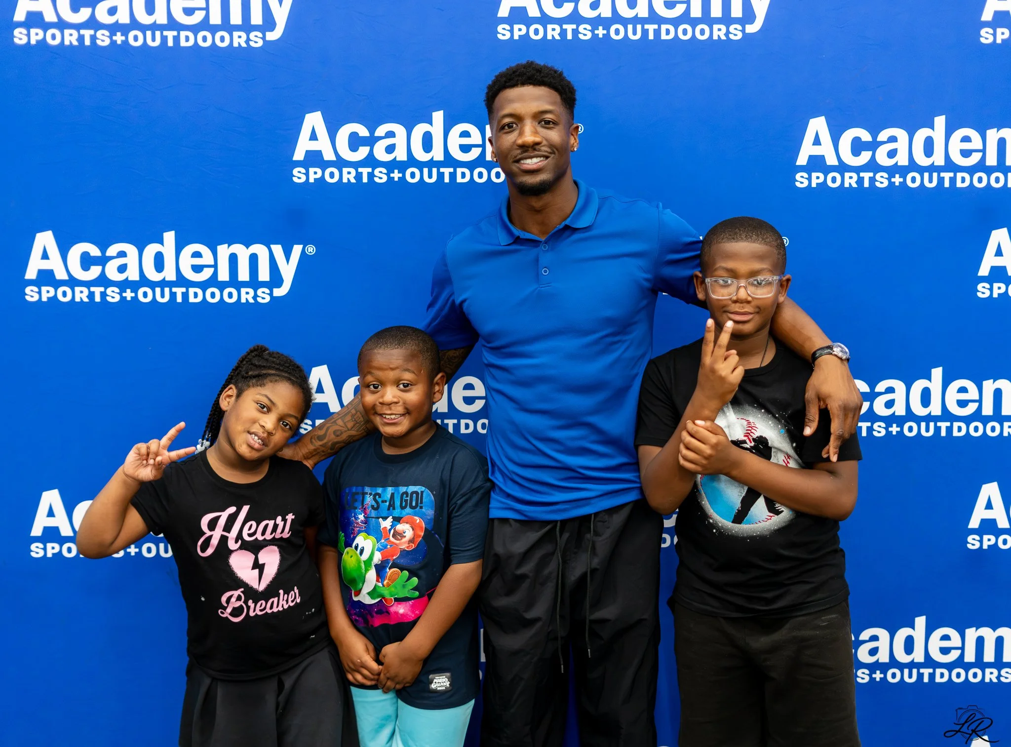 A man and four children posing together in front of a blue backdrop with 'Academy Sports + Outdoors' logos. The man is wearing a blue polo shirt, and the children are wearing casual t-shirts. The children are making peace signs and smiling.