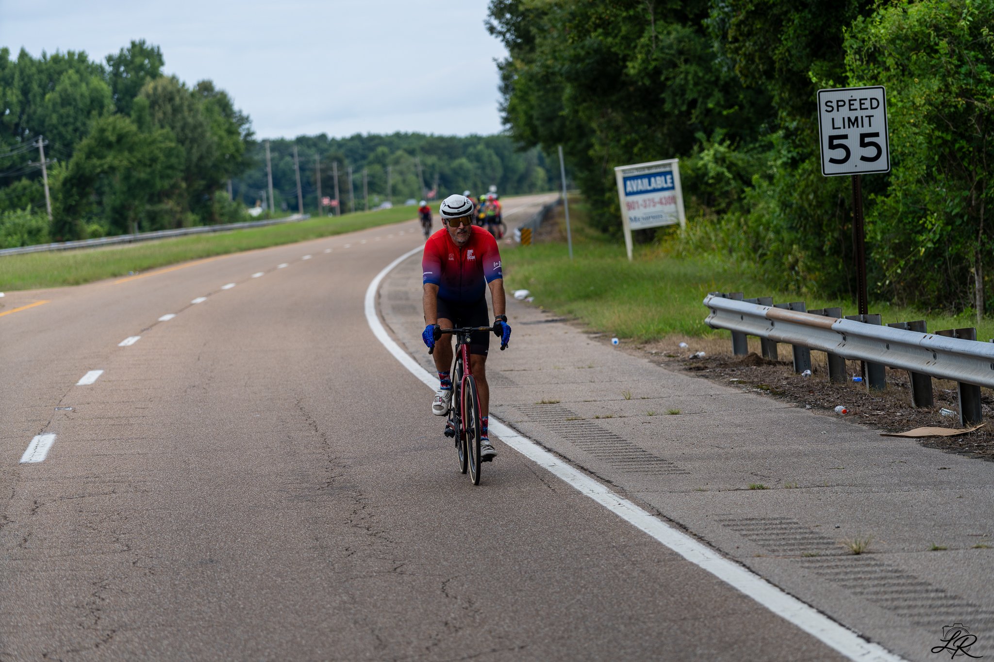 A person wearing a red and blue cycling jersey, black shorts, and a white helmet cycling on the side of a curved road surrounded by trees and greenery. Several other cyclists are visible in the background. A speed limit sign indicates 55 mph, and the