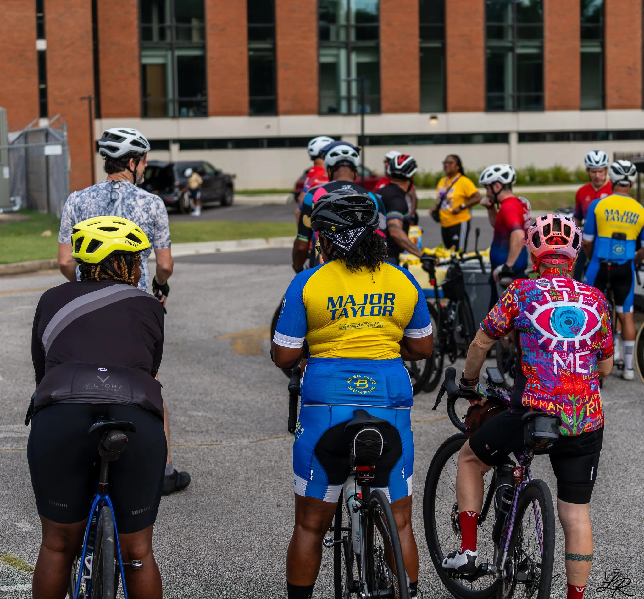A group of cyclists gathered outdoors, wearing helmets and sports attire, with a building in the background.