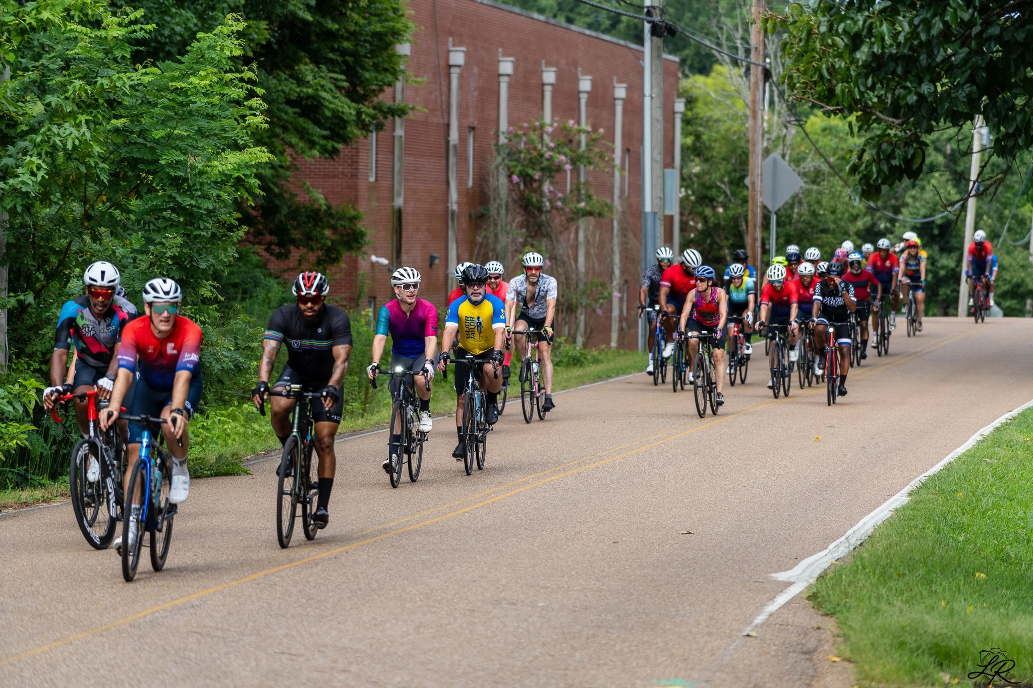 Group of cyclists riding on a paved road next to a brick building and green trees.