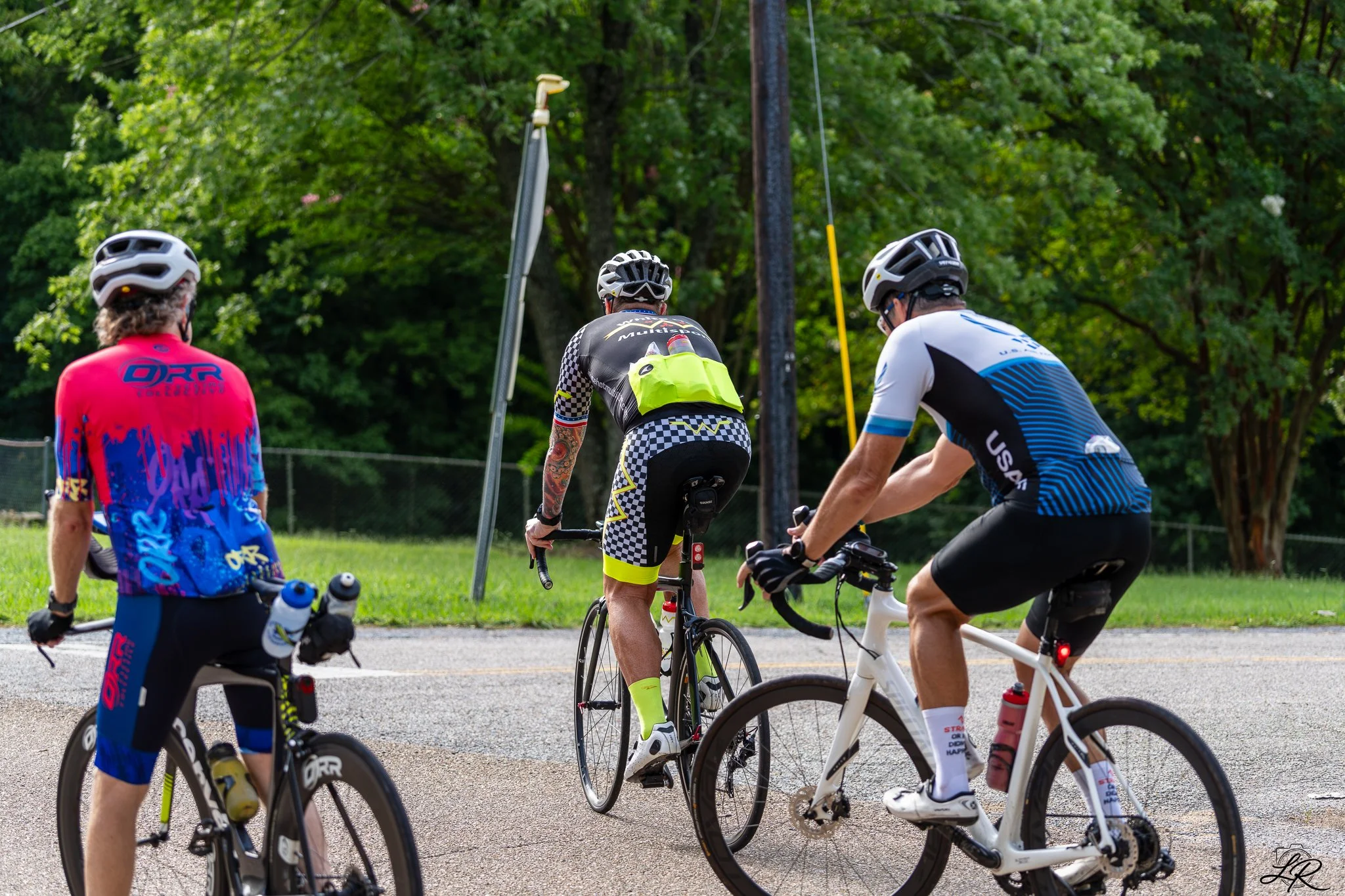 A group of cyclists gathered outdoors on a paved path, with lush green trees in the background. Some riders wear helmets and sports clothing, and they appear to be preparing for or taking a break from cycling.