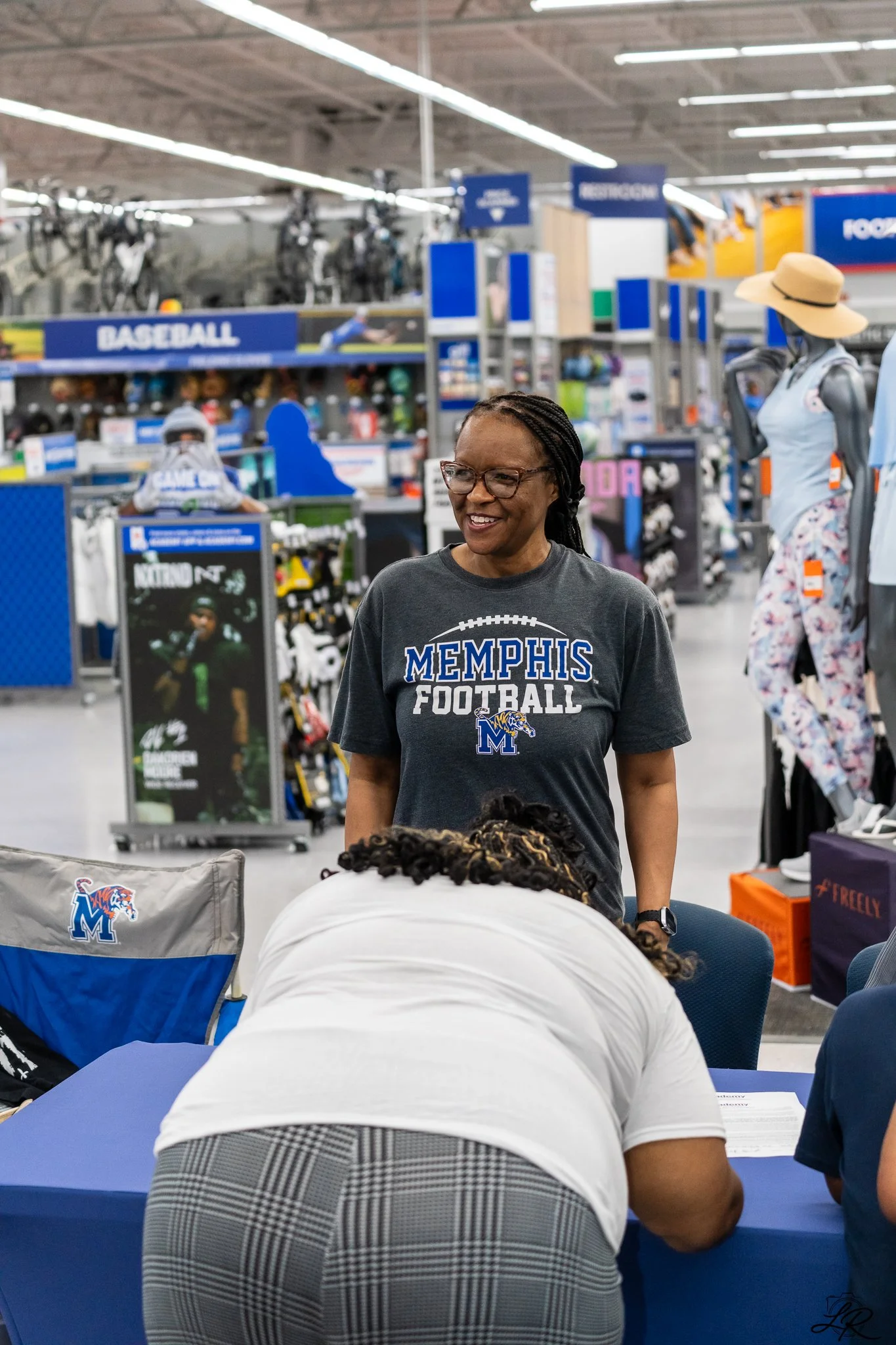 A woman wearing glasses and a Memphis Football shirt smiling while talking to a group of people in a store, with shelves filled with sporting goods and mannequins dressed in sports attire in the background.