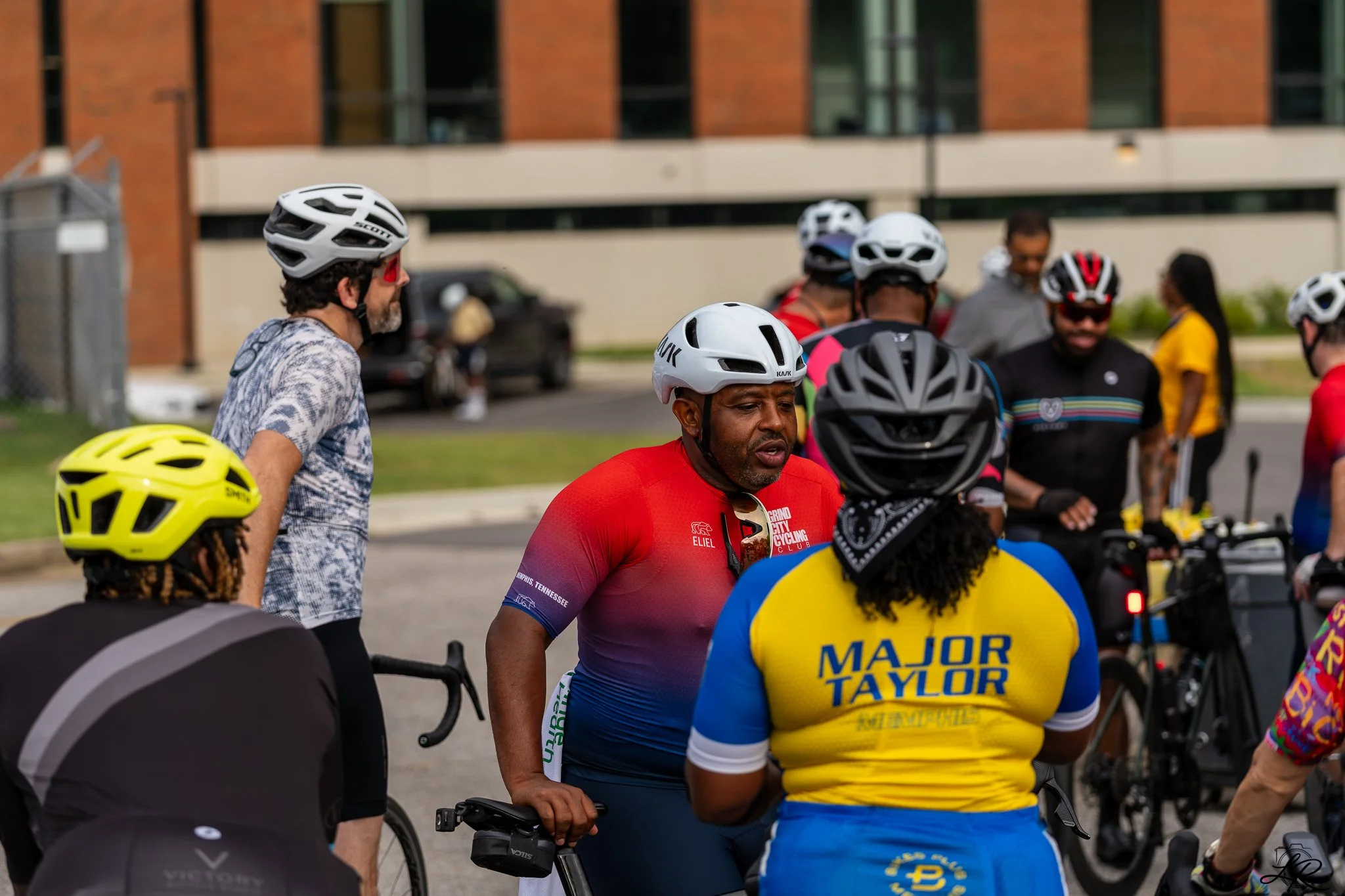 A group of cyclists gathered outdoors, wearing helmets and cycling gear, near a building on a city street.