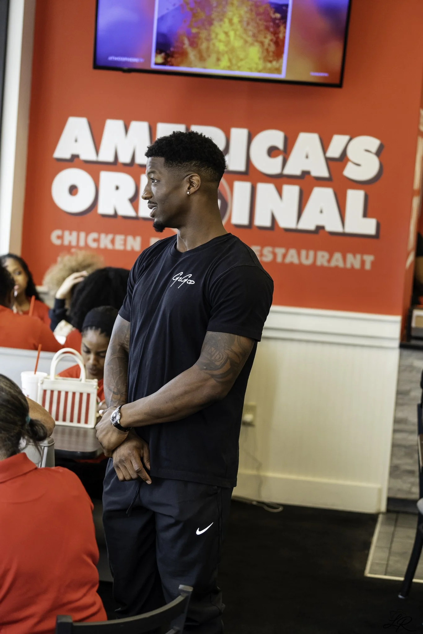 A young man with short curly hair, earrings, and tattoos on his forearms is standing inside a restaurant. He is wearing a black t-shirt with a white logo on it. Behind him is a red wall with large white text that says 'AMERICAS ORIGINAL' and smaller 