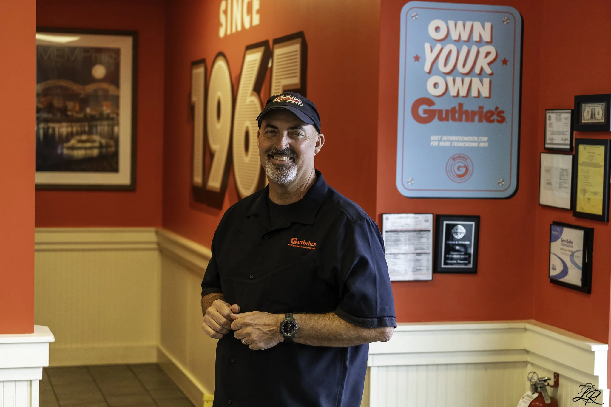 A man wearing a black shirt and cap with a Guthrie's logo, smiling inside a restaurant with red walls and signs, including one that reads 'Own Your Own Guthrie's'.