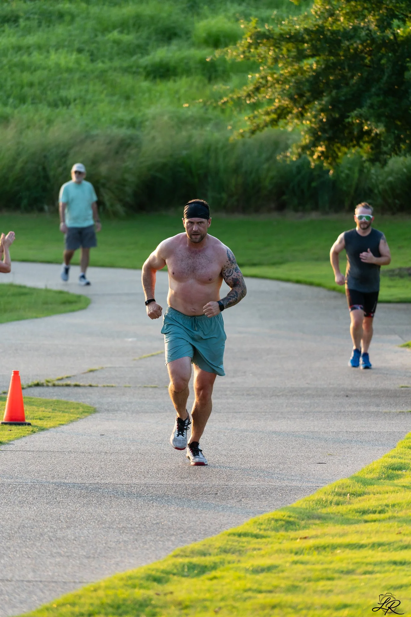 A group of men running on a paved path in a park during sunset. One man is shirtless, wearing a headband and shorts. Others are dressed in t-shirts and shorts or pants. There are orange cones marking the path, and trees and grass surround the area.