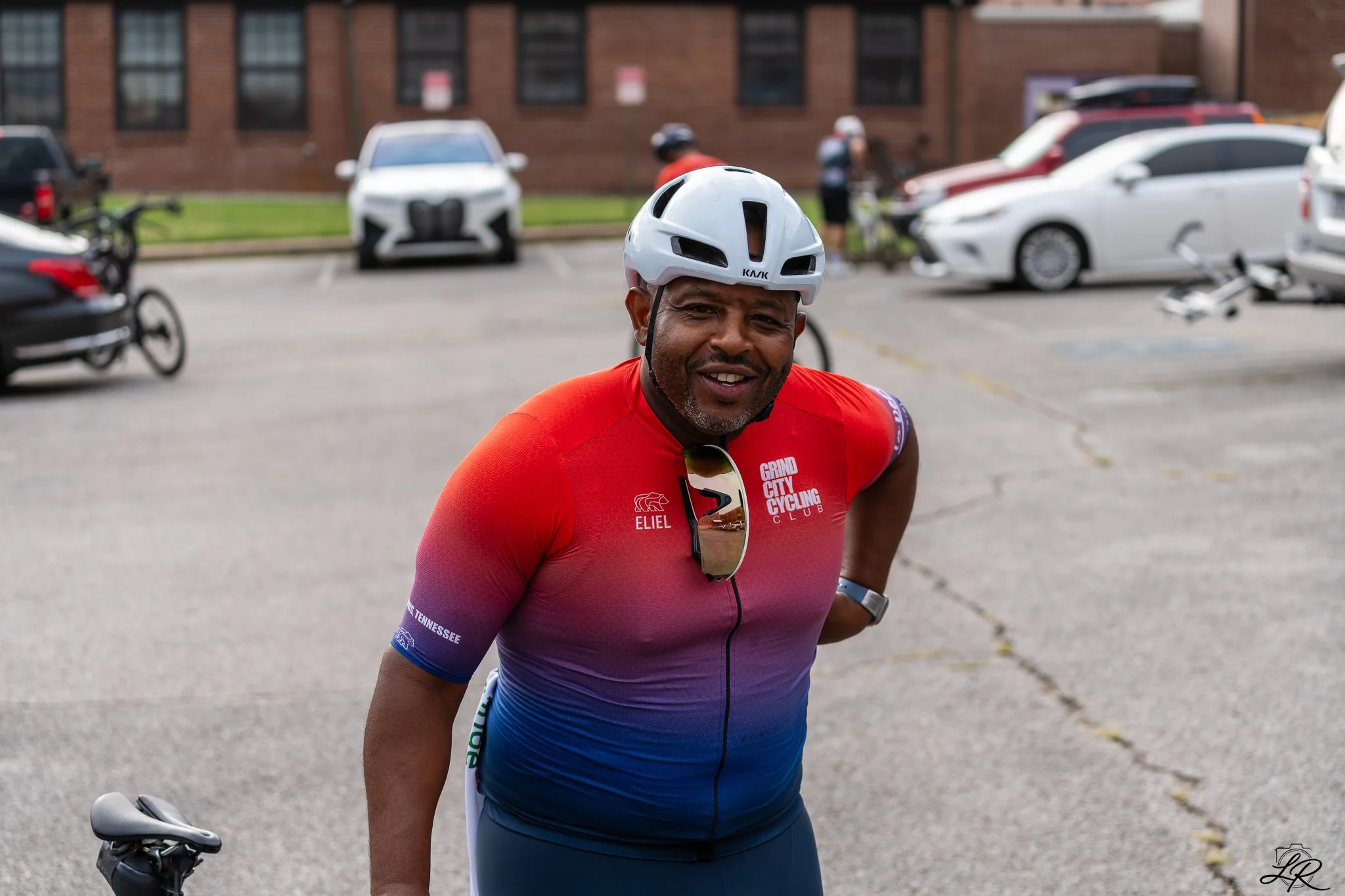 A man wearing a white cycling helmet and a red and purple cycling jersey standing in a parking lot with parked cars and bicycles in the background.