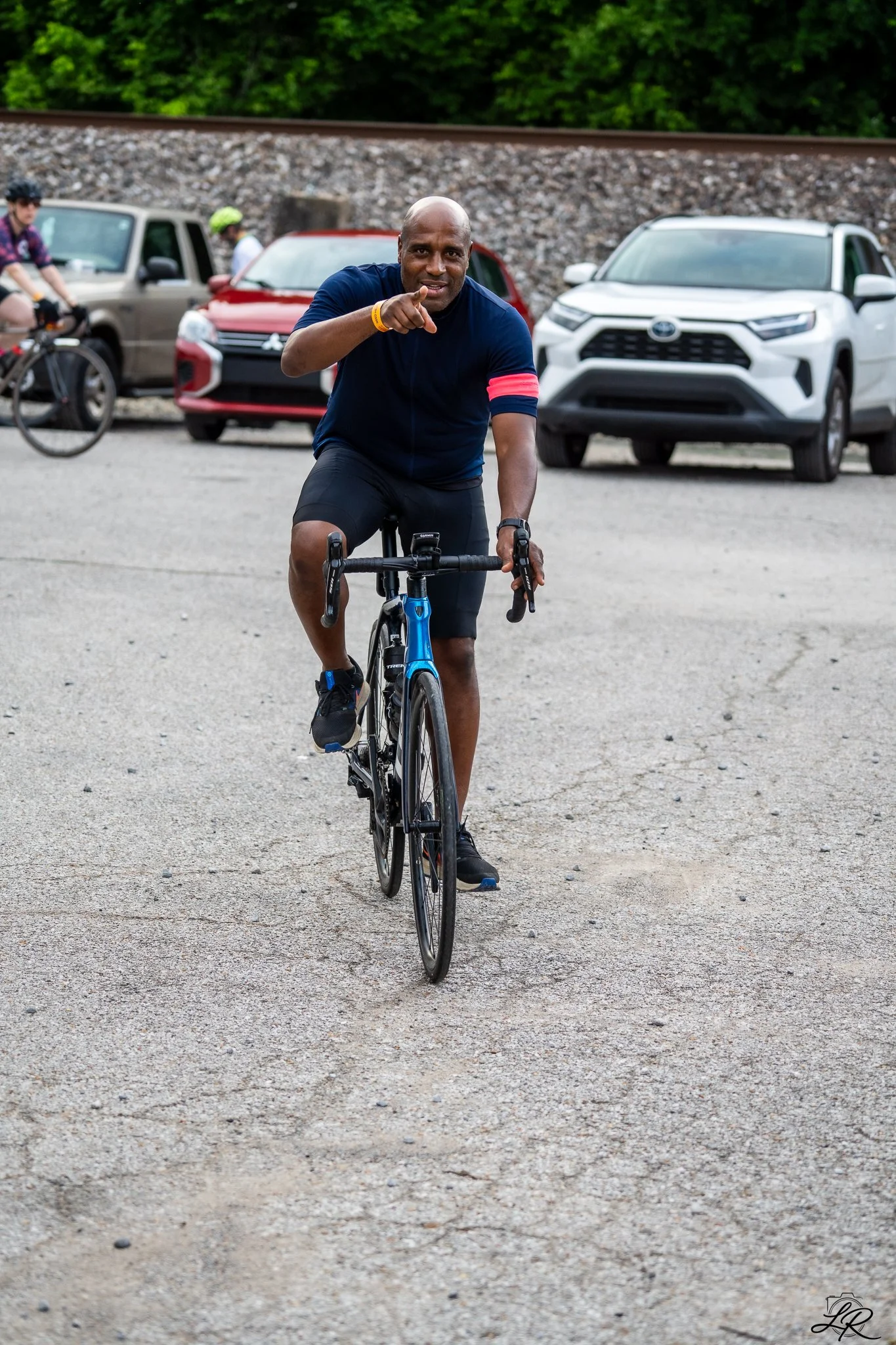 A man in a dark blue cycling jersey and black shorts is riding a road bike on a gravel surface, pointing forward, with cars and other cyclists in the background and green foliage behind a railroad track.