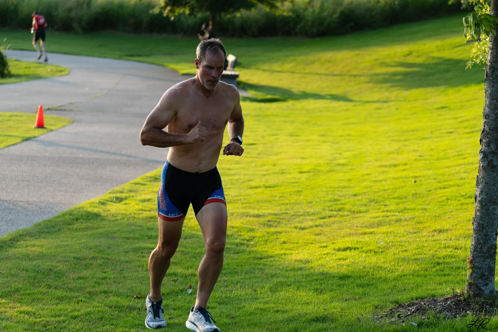 A shirtless man jogging on a paved path in a park with green grass and trees, with another person visible in the background.