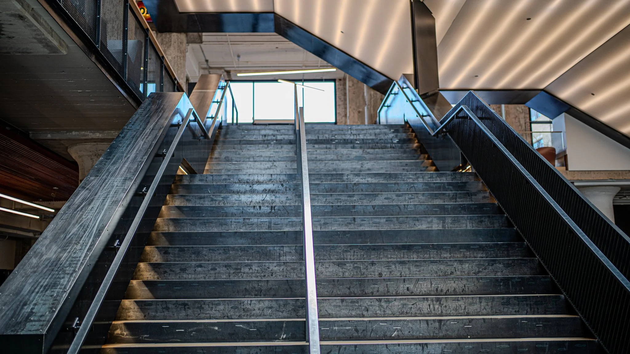 Indoor modern staircase with black metal railings and steps, overlooking a spacious area with large windows and contemporary design elements.