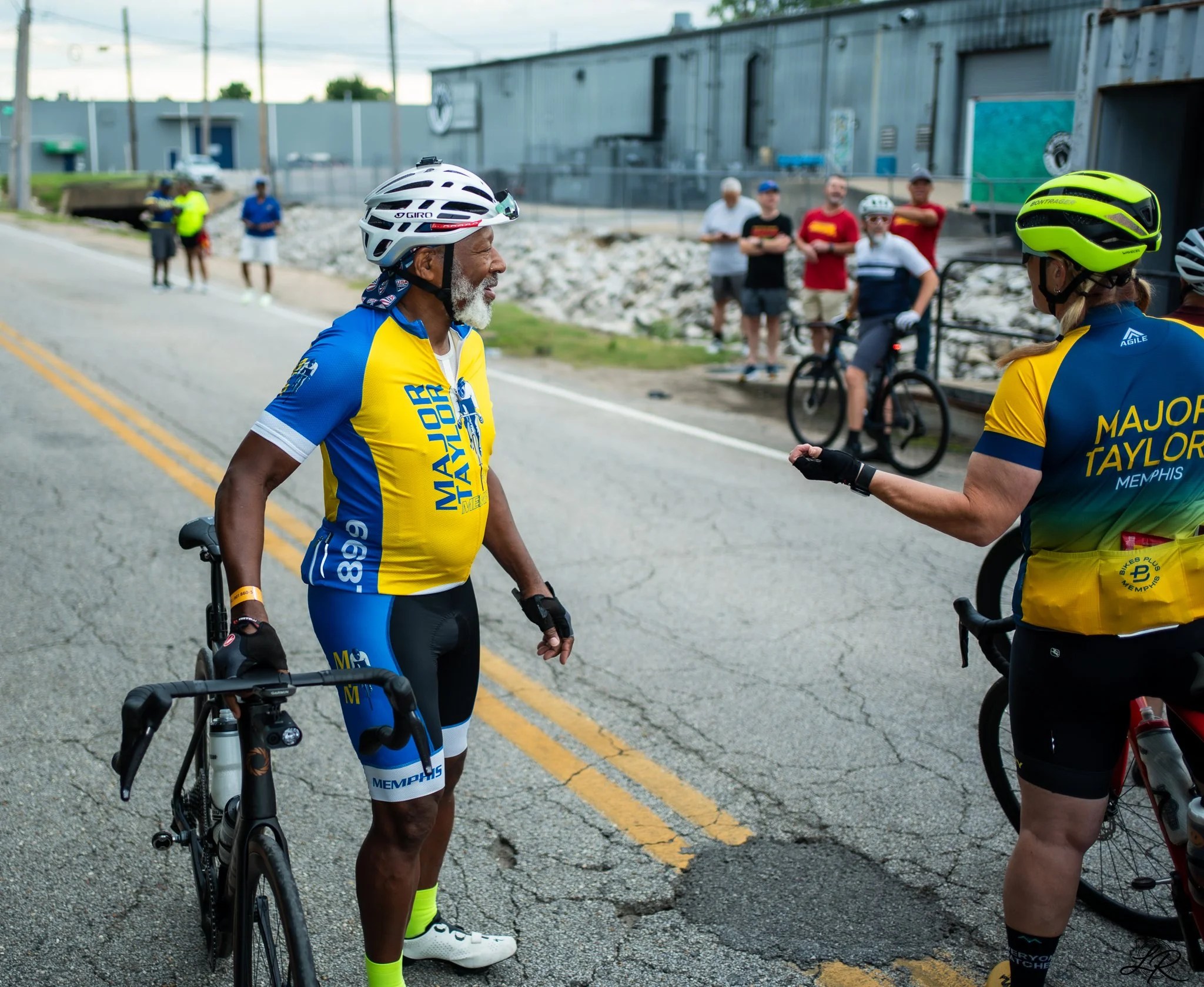 A group of cyclists in colorful jerseys and helmets socializing on a cracked street, with a metal industrial building and some onlookers in the background.