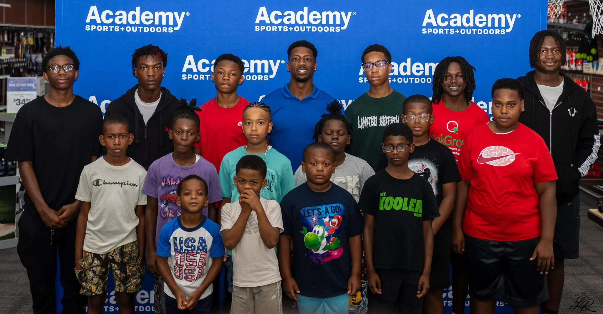 Group of children and young adults posing for a photo in front of a blue backdrop with 'Academy Sports + Outdoors' logo in a store.