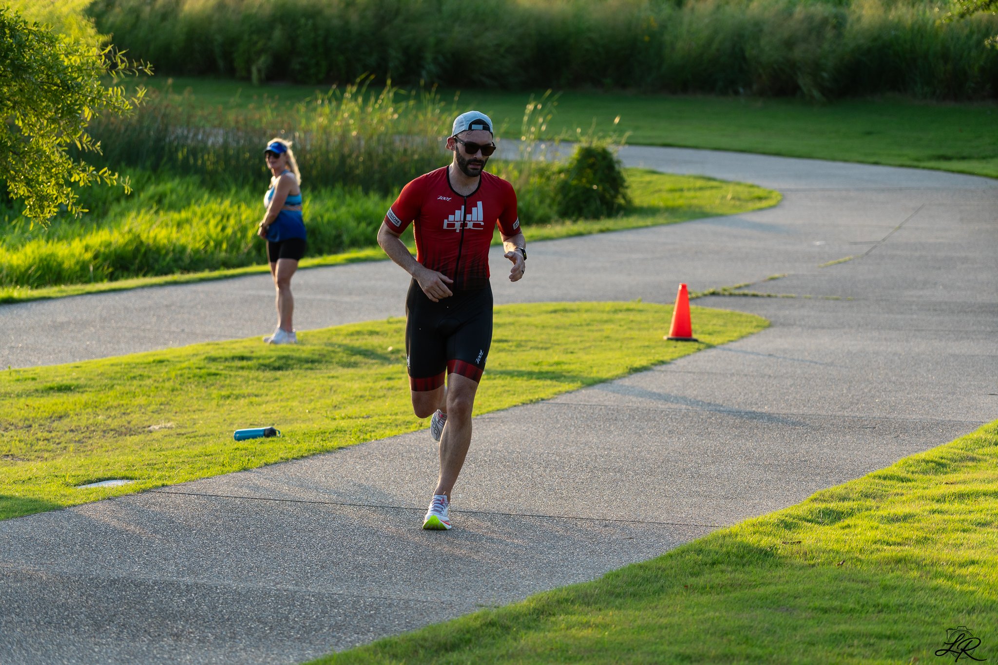 A man running outdoors on a paved and grassy trail, wearing a red athletic shirt, black shorts, sunglasses, a cap, and a watch, with a woman walking behind him on a sunny day with green trees and a pond in the background.