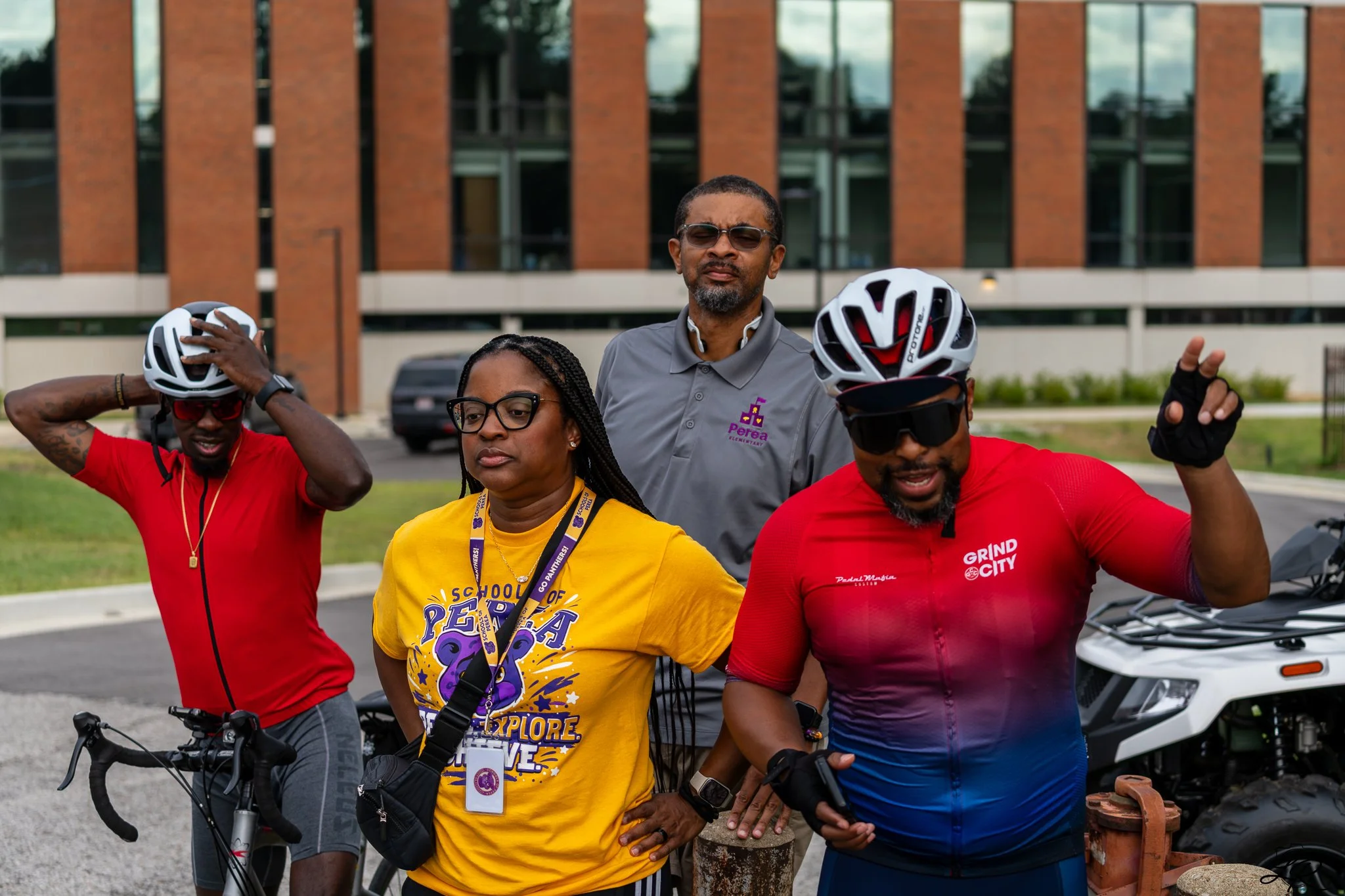 A group of four diverse people in athletic clothing and helmets, standing outdoors in front of a brick building and parking lot, preparing for a biking event.