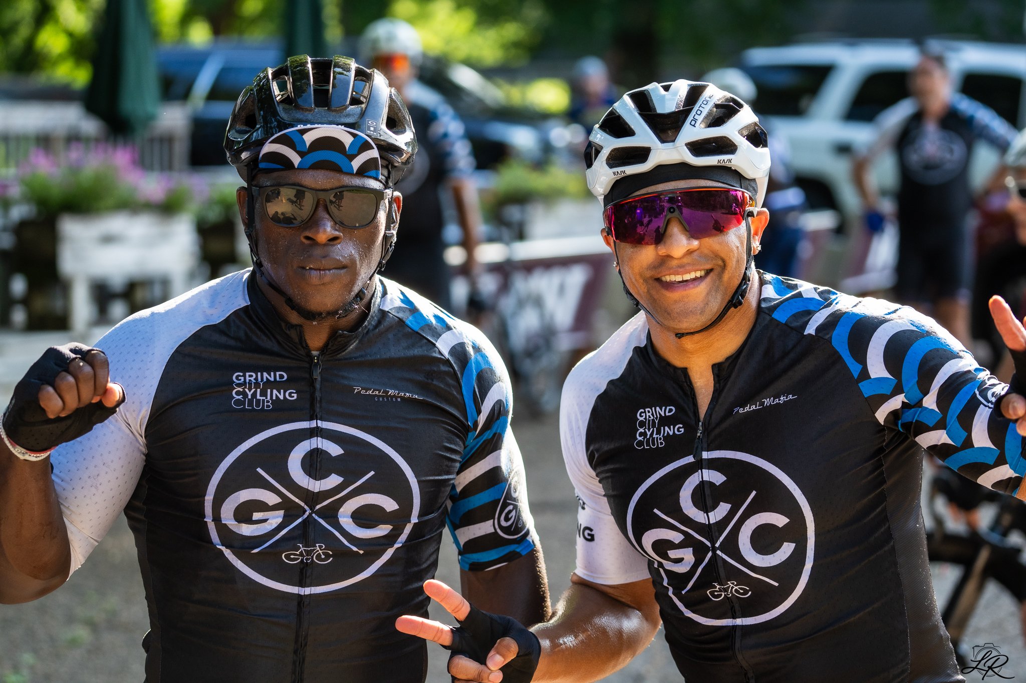 Two cyclists in cycling jerseys and helmets posing for a photo outdoors, with trees and cars in the background.
