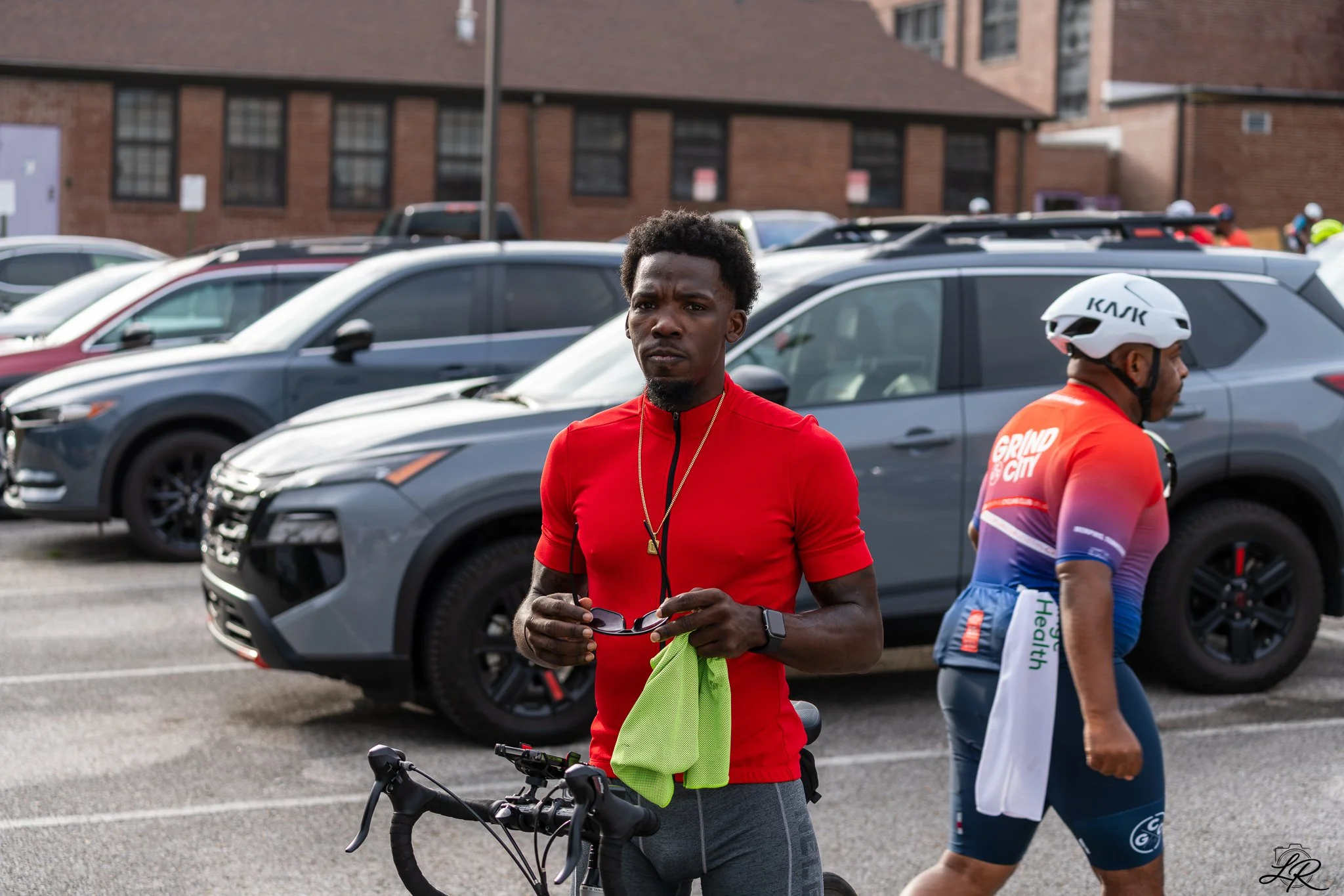 A young man with dark curly hair, wearing a red athletic shirt, holding a green cloth and glasses, standing next to a bicycle in a parking lot. Another cyclist wearing a helmet and a colorful cycling jersey is partially visible beside him. Cars and a