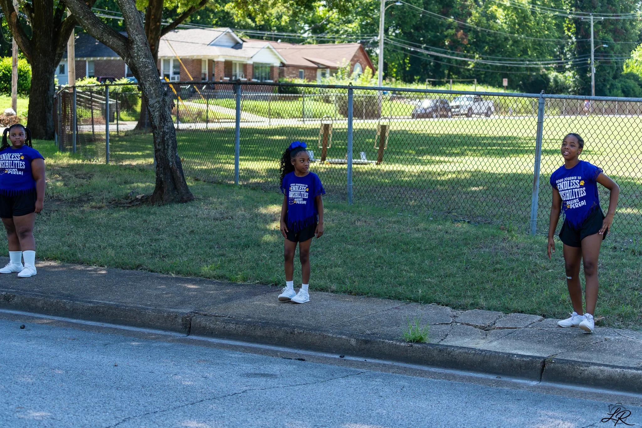 Three young girls are standing outside near a chain-link fence, in a suburban neighborhood on a sunny day. They are wearing matching blue t-shirts and black shorts.