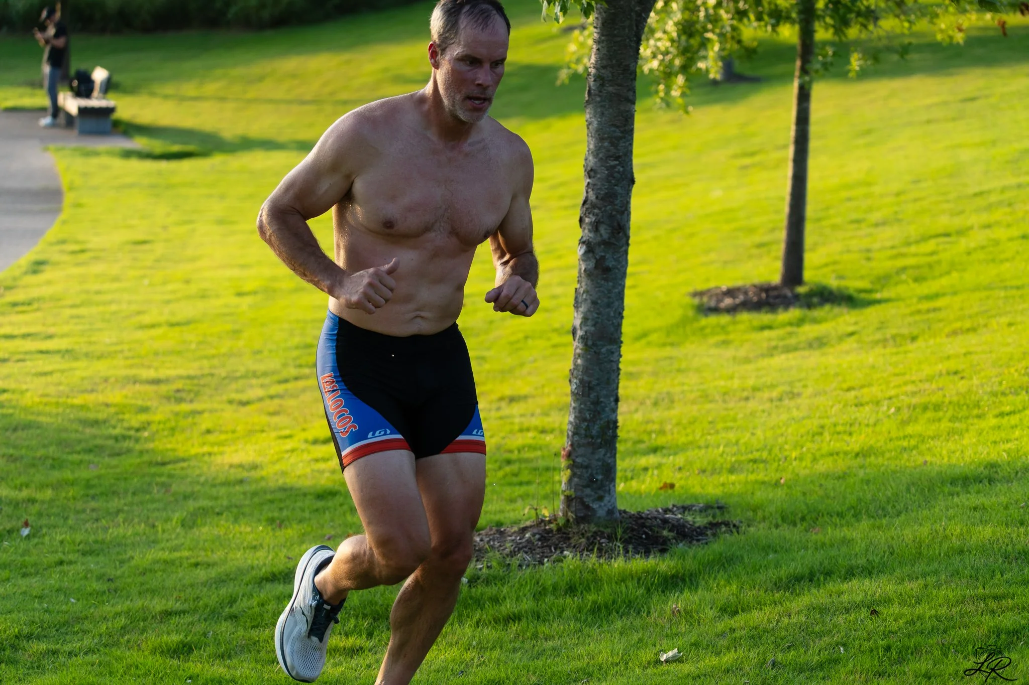 A shirtless man running outdoors in a park, with trees and a person in the background taking photos.