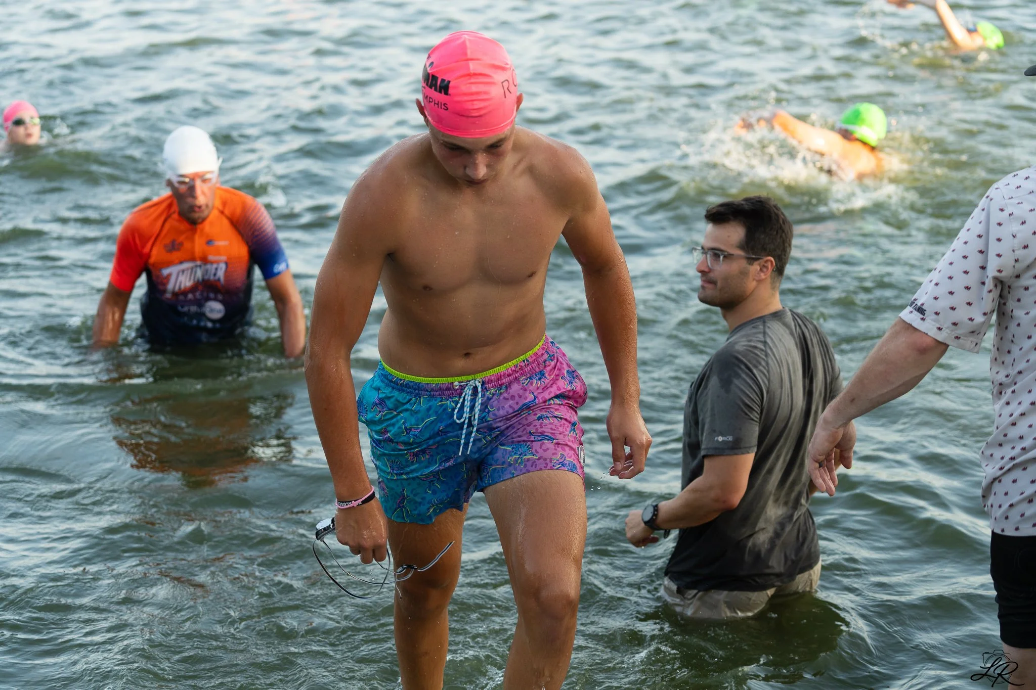 A group of people at the beach with some in wetsuits and swimming caps, some standing in the water, getting ready for a swimming event or training session.