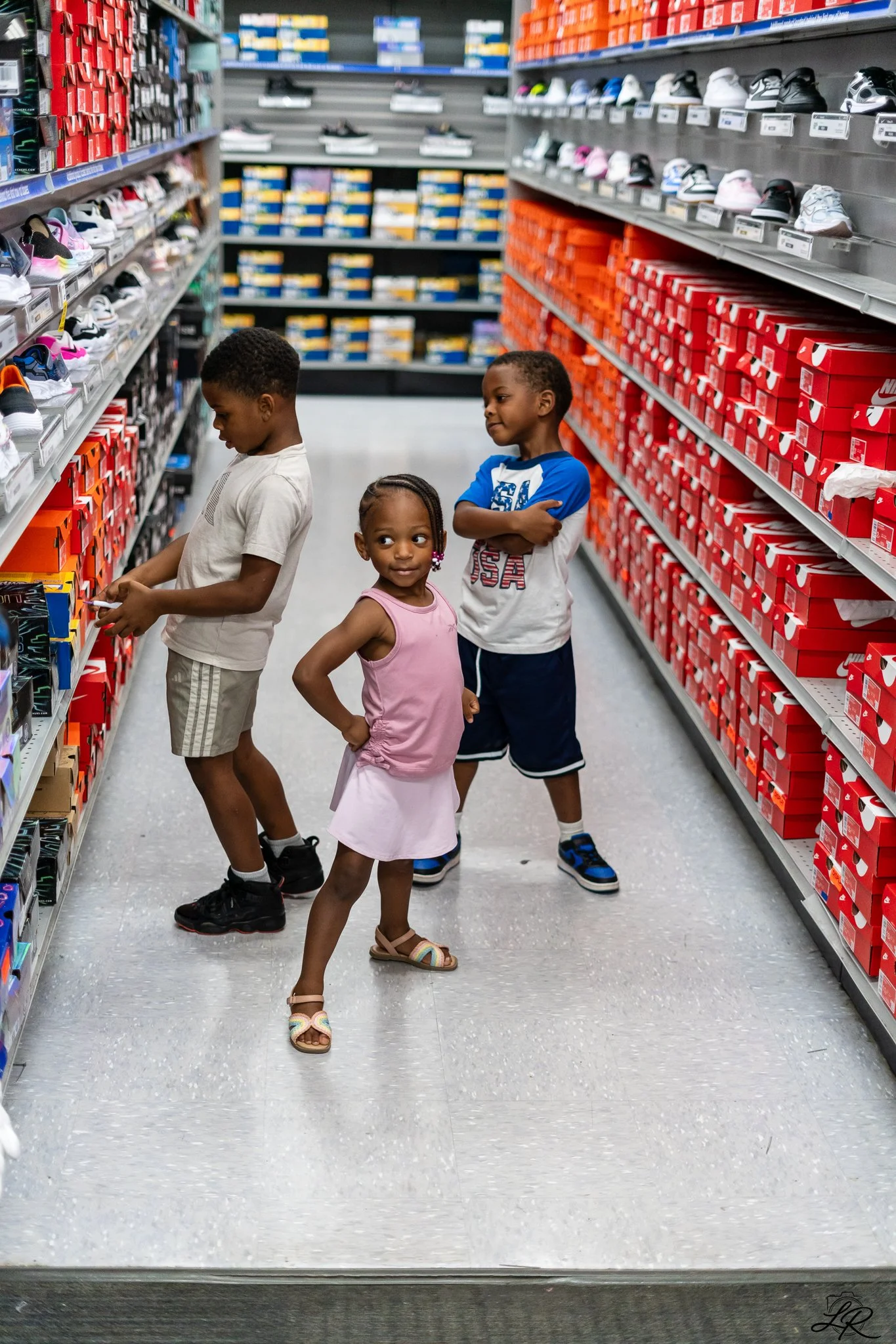 Three children shopping for shoes in a store, standing between shelves filled with various athletic shoes and shoeboxes.
